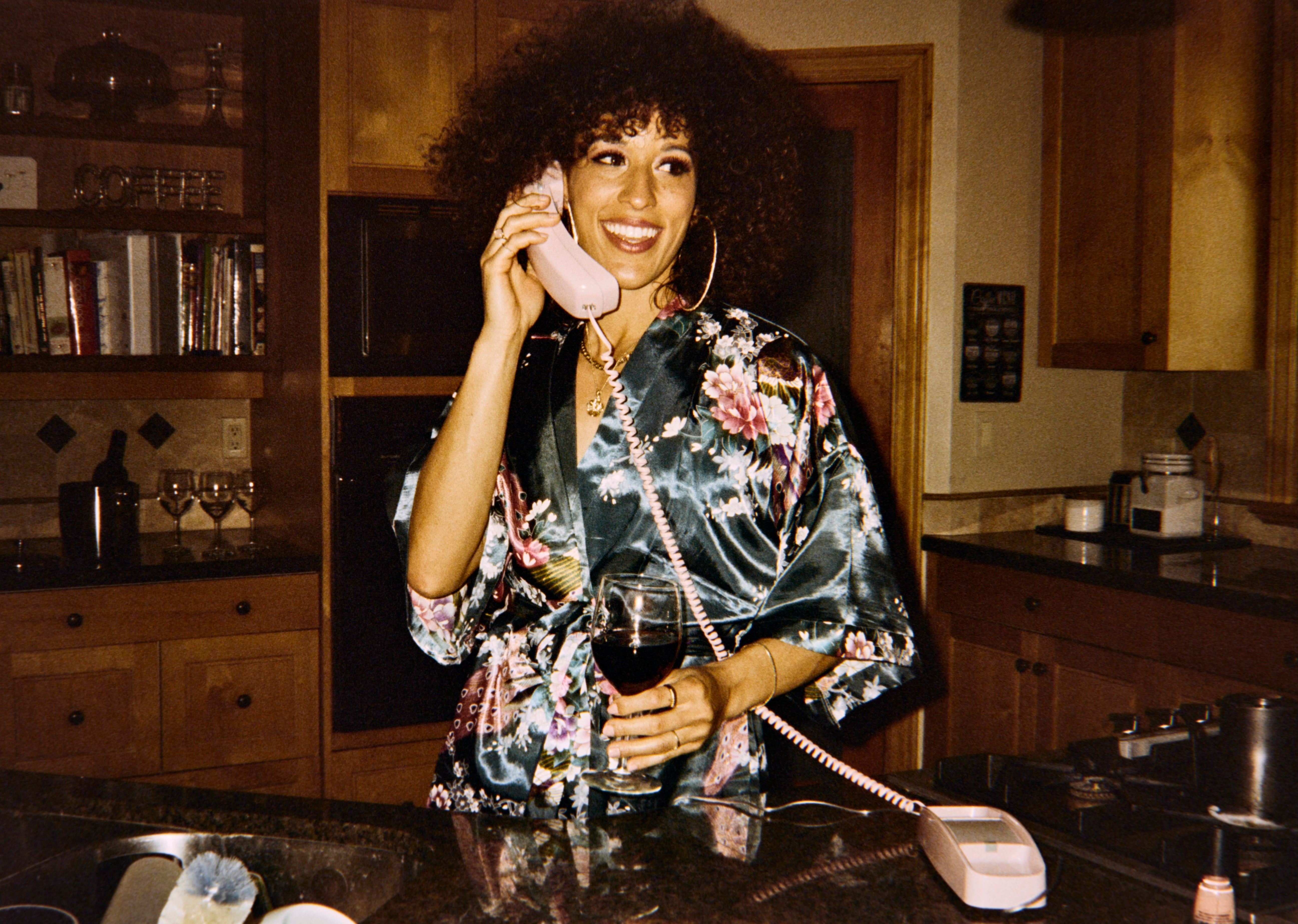 Person in floral robe, holding a glass of wine, talks on a vintage corded phone in a cozy kitchen setting, evoking retro vibes