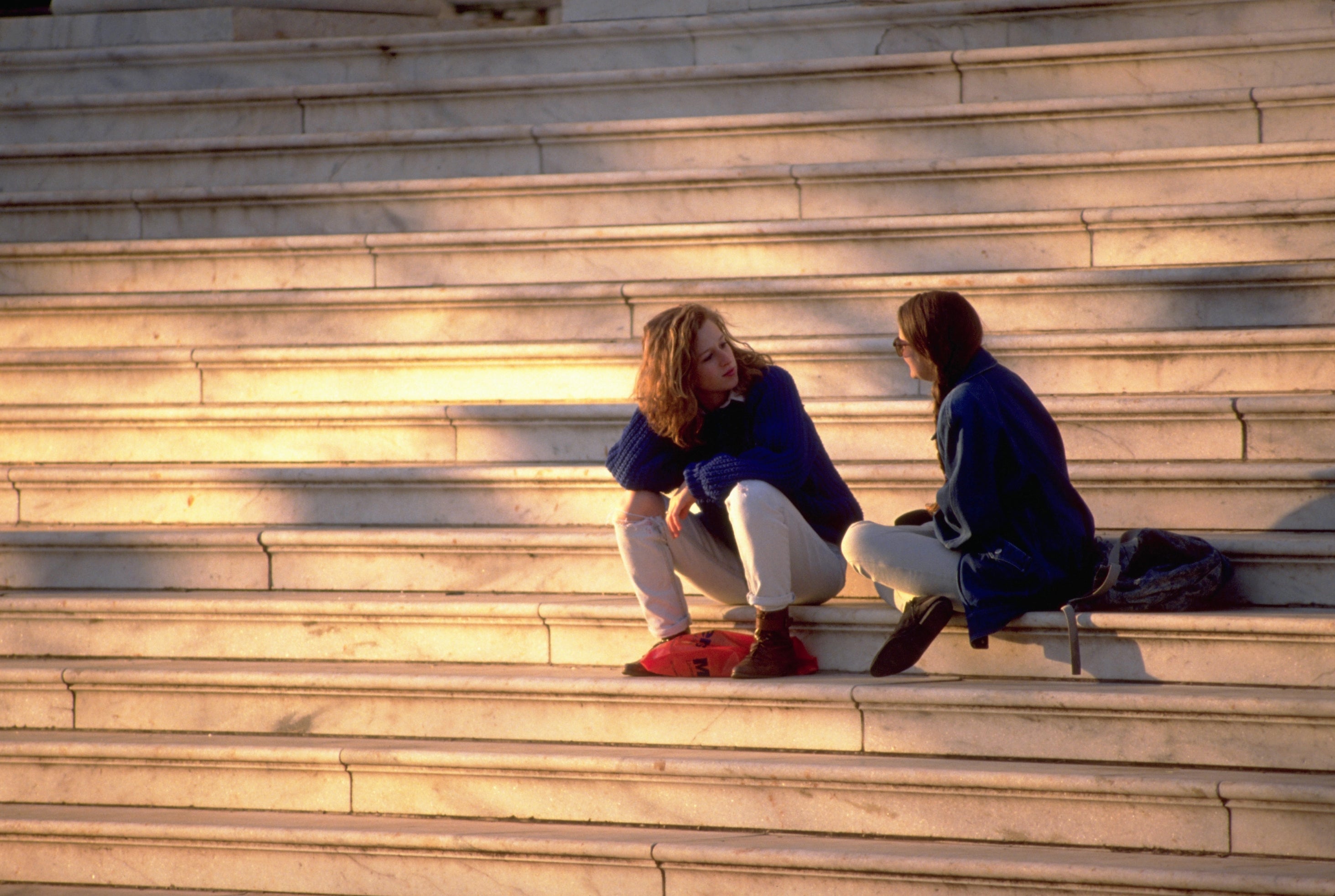 Two people sit talking on outdoor stone steps, both wearing casual clothing