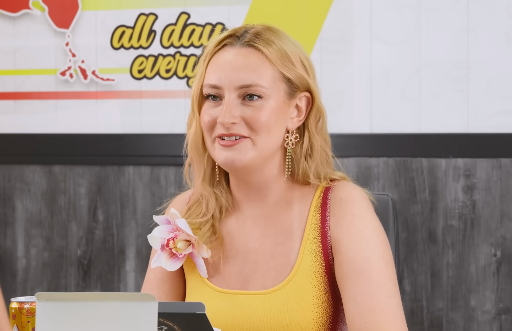 A person sitting at a table, wearing a sleeveless top with floral decoration. They have long hair and are smiling. A design and text are in the background