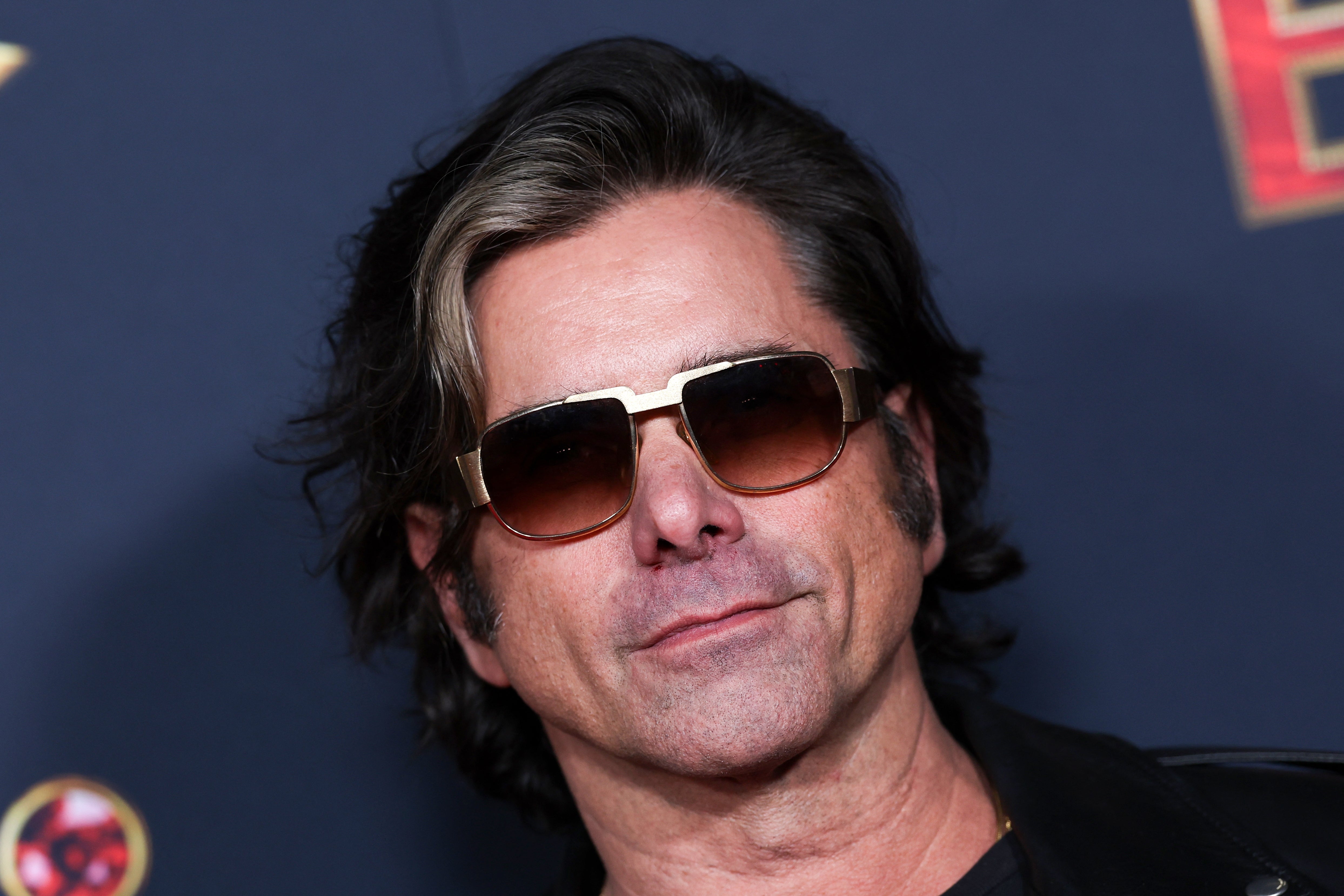 A person with long hair wearing sunglasses and a jacket poses confidently at a formal event