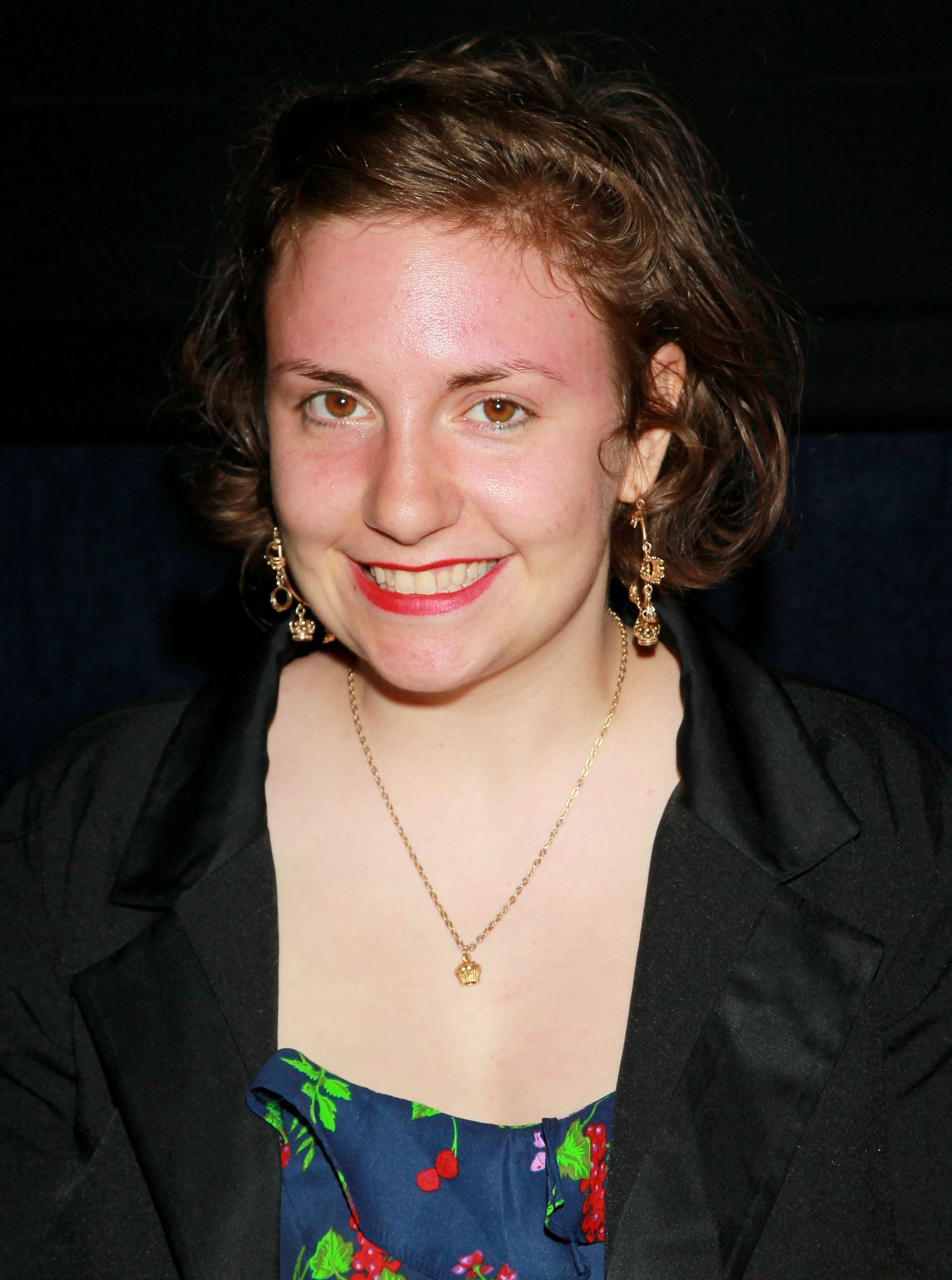 Person with short, tousled hair wearing a dark jacket, floral top, and gold necklace, smiling at an event