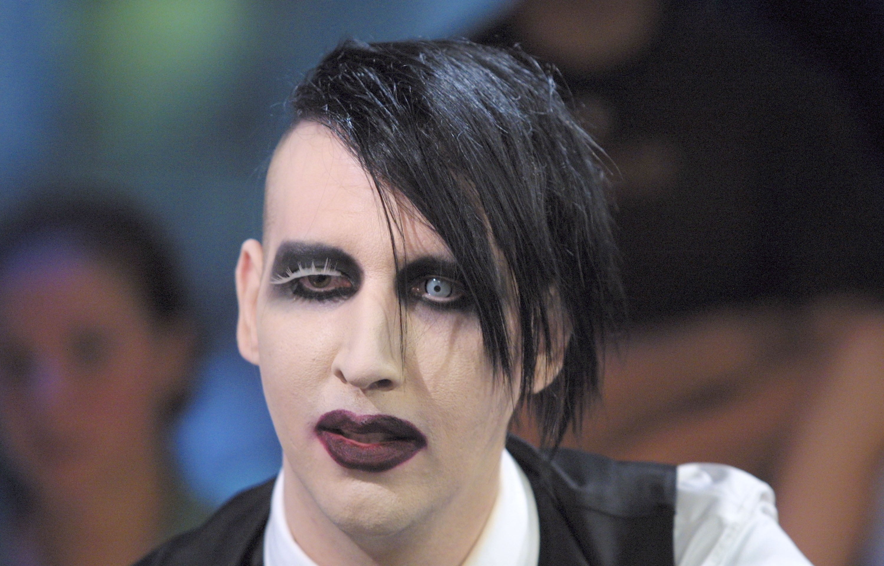 Person with gothic-style makeup, striking hairstyle, wearing a dark vest over a white shirt