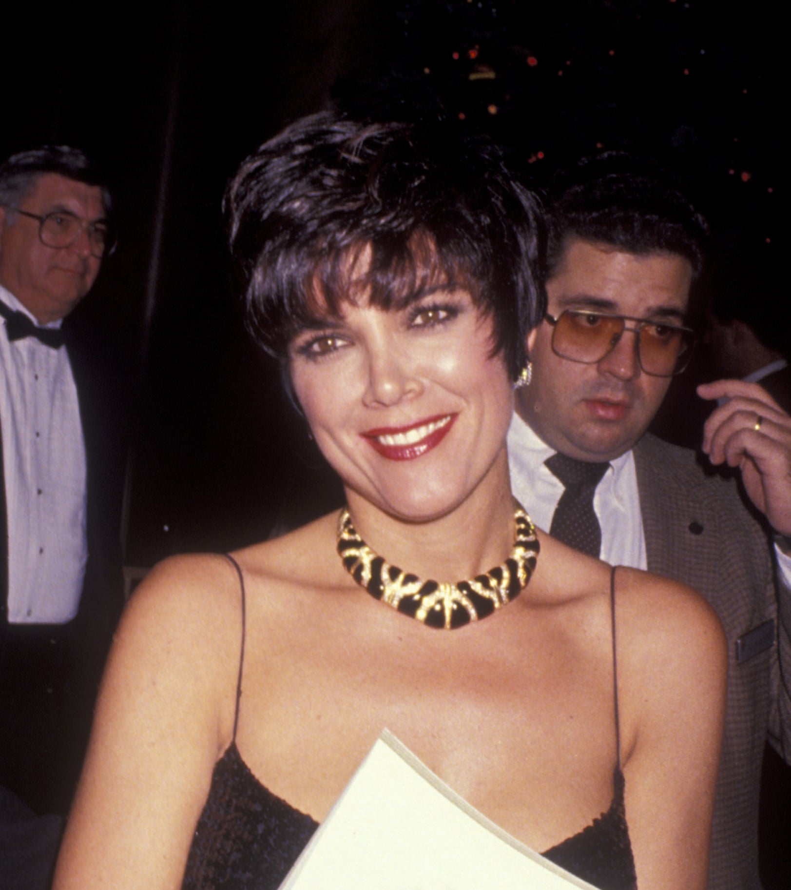 Person in a black sequin dress holds a book with a man's photo on the cover, smiling at an event
