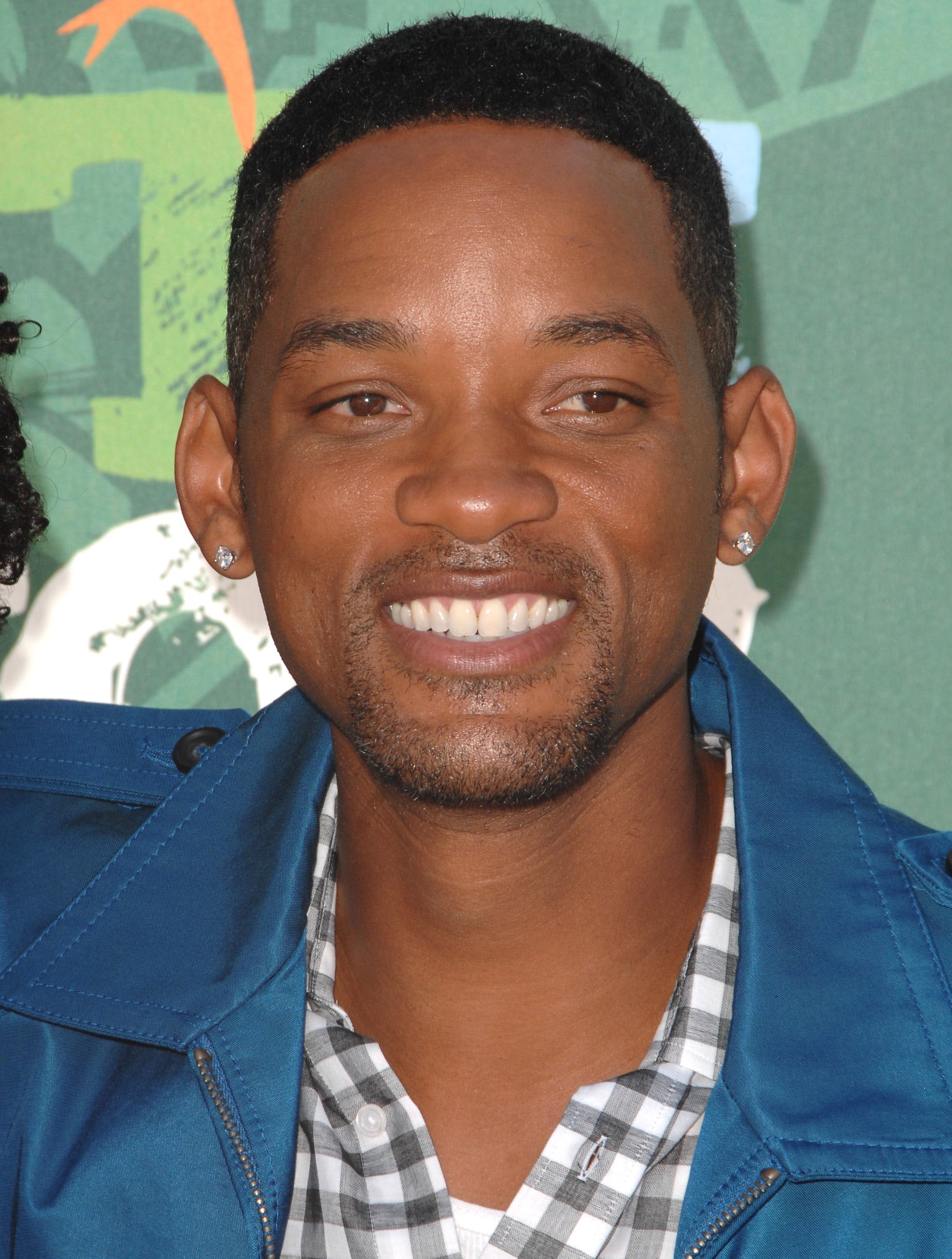 A person smiles for a photo, wearing a checked shirt and a blue jacket