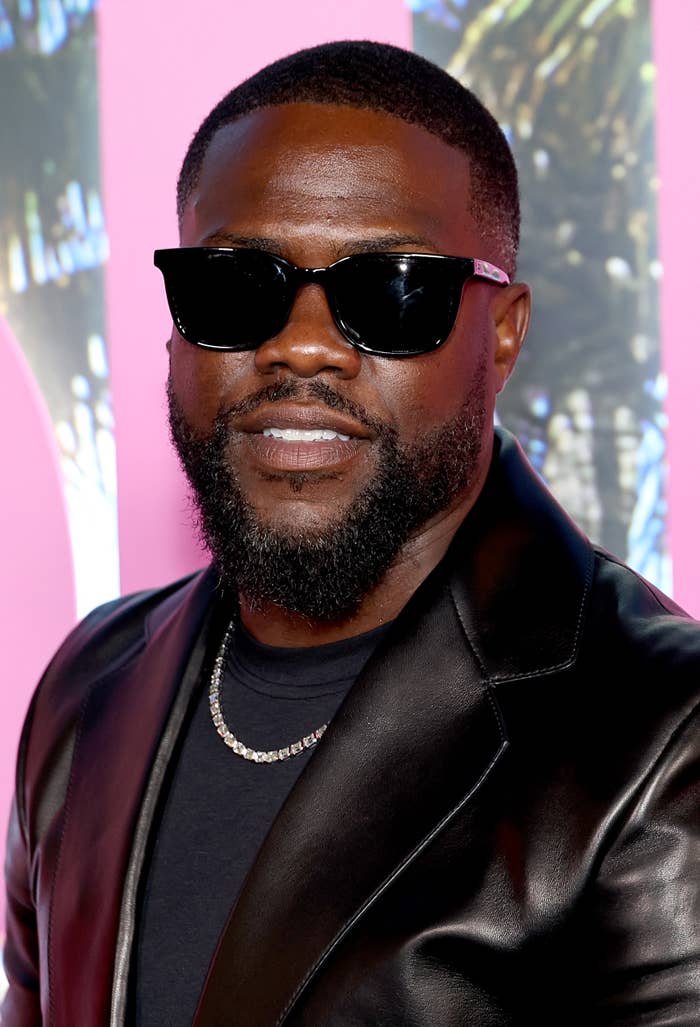 Man in sunglasses wearing a sleek black leather jacket and a necklace, smiling at an event