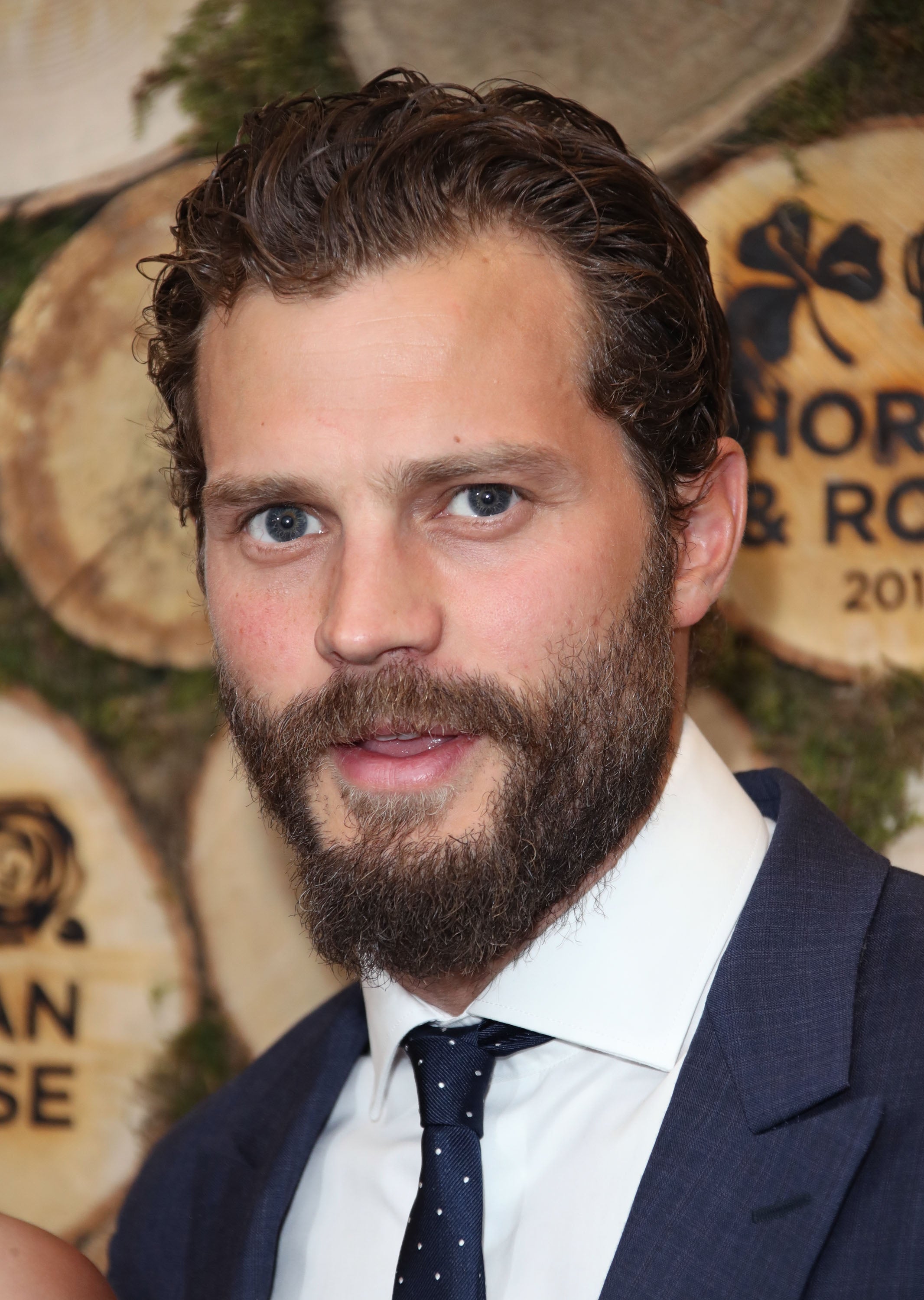 Bearded man in a suit and polka-dot tie at an event, with a background of circular wooden plaques