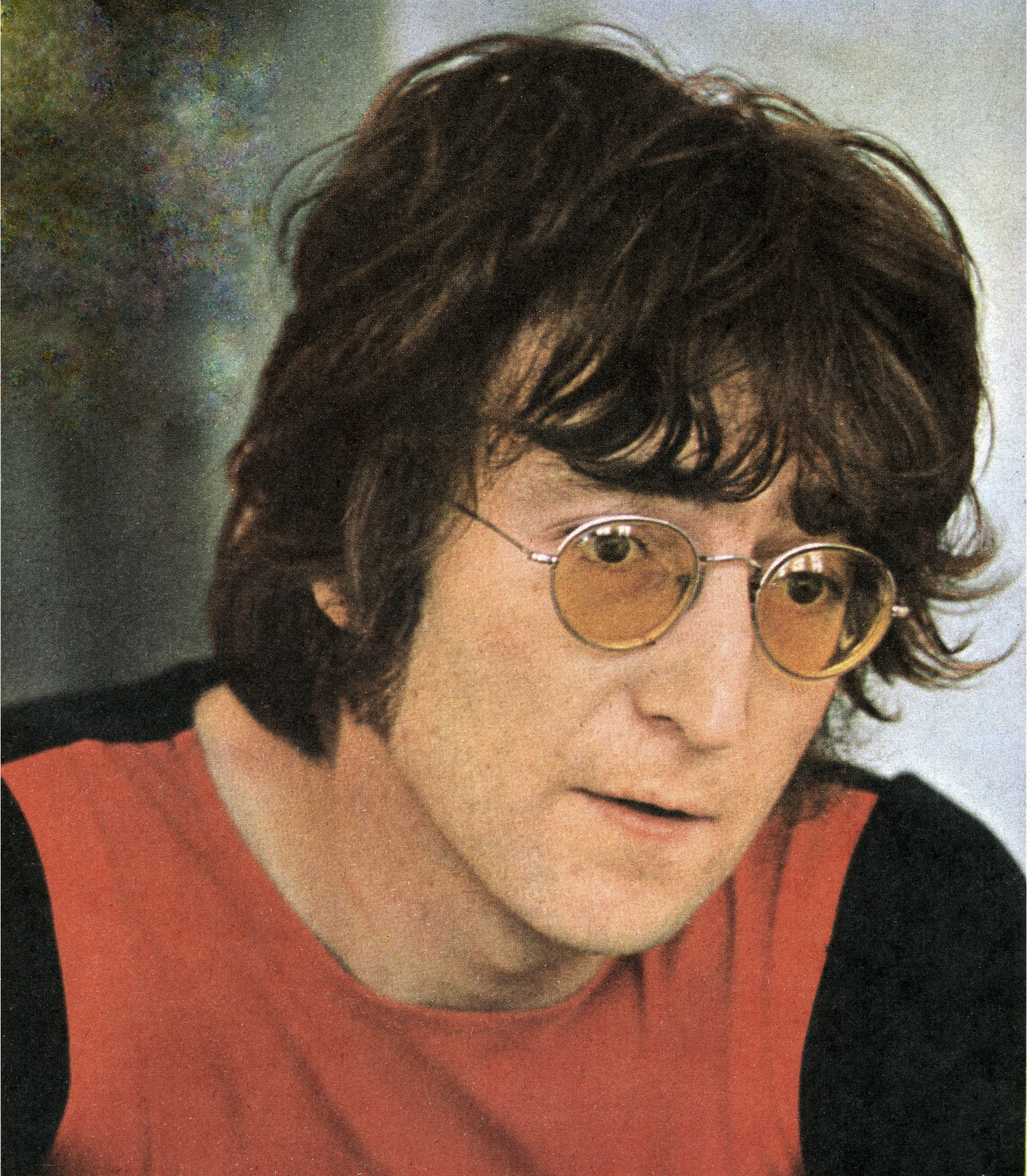 A person with wavy hair and round glasses is facing forward, wearing a casual outfit with a serious expression
