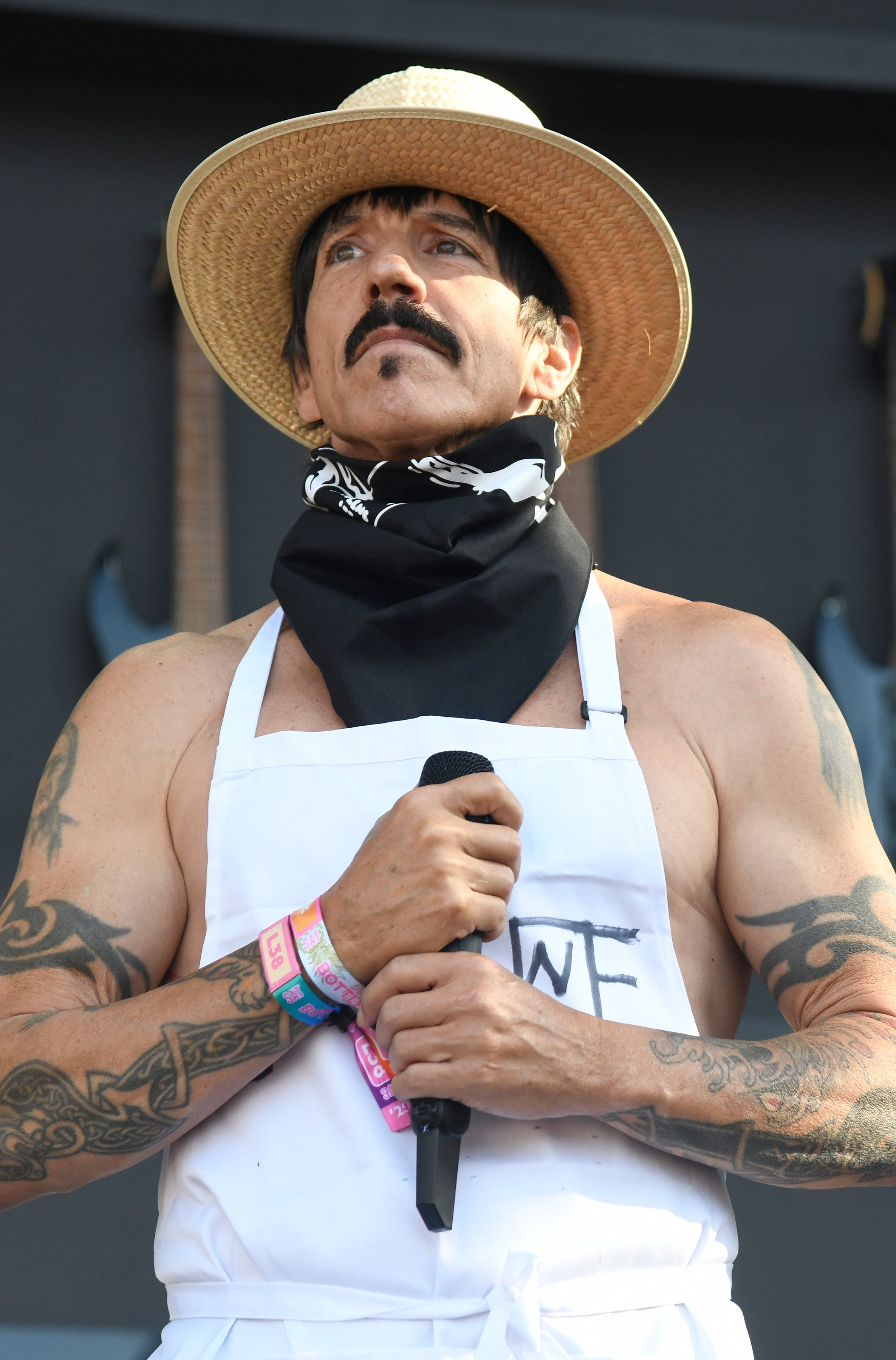 A man wearing a straw hat, a bandana, and an apron holds a microphone at an outdoor event