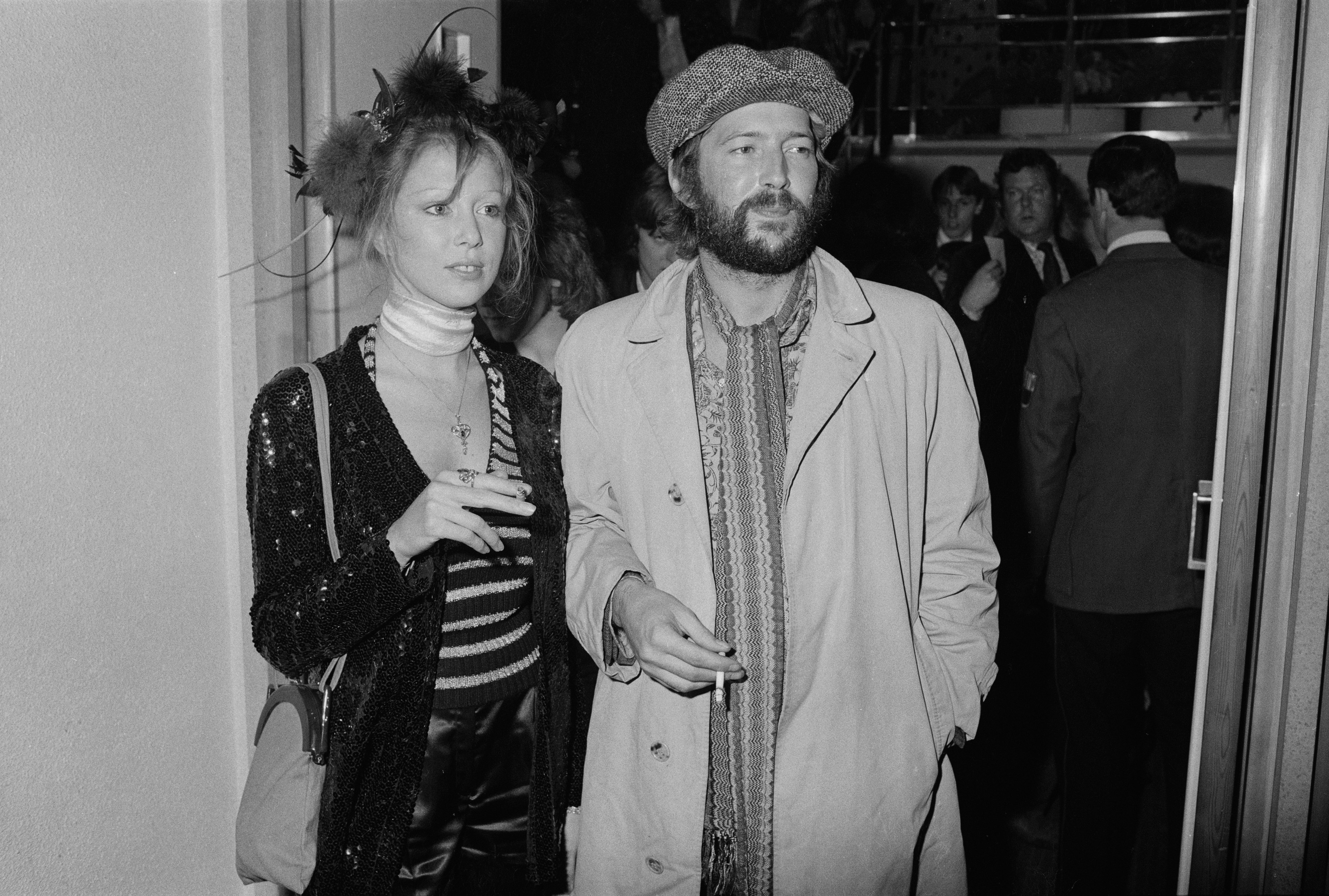 A woman in a striped top with a sparkly jacket and a man in a trench coat and beret hold cigarettes at an event with people in the background