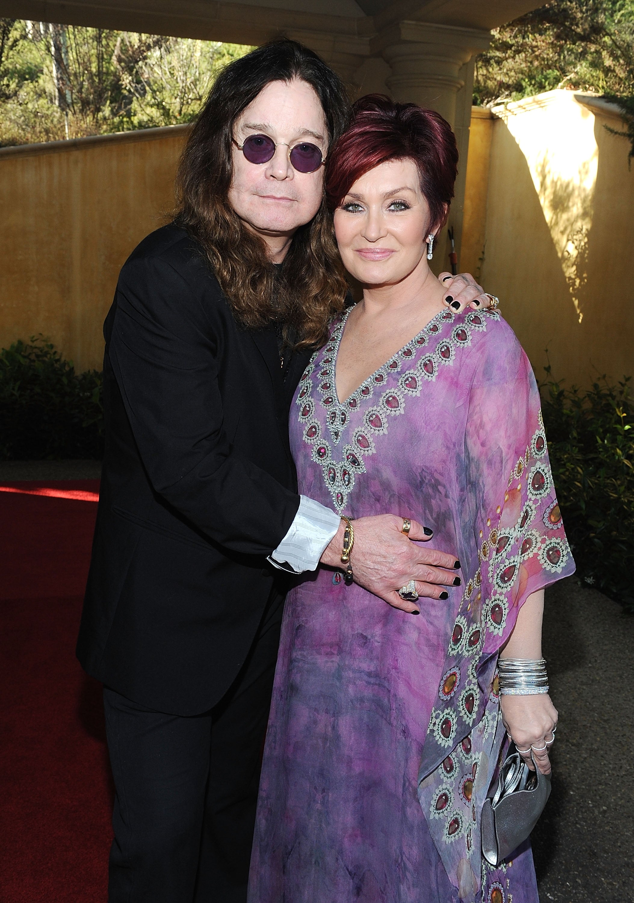 Two individuals pose on a red carpet. One wears a dark suit and round glasses, and the other wears a flowing gown with intricate designs