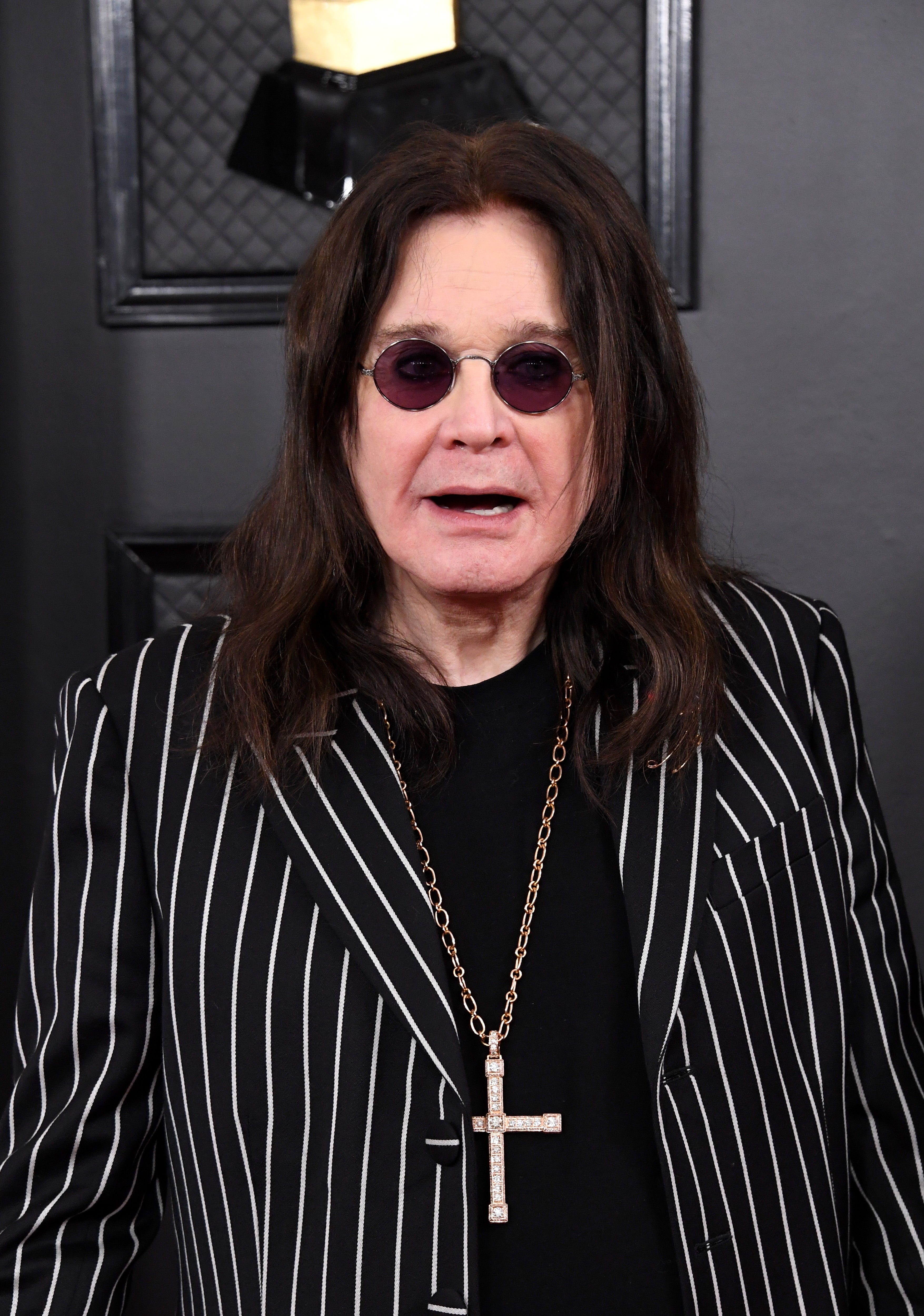 Person with long hair, wearing round sunglasses, striped blazer, and cross necklace, standing in front of an award backdrop
