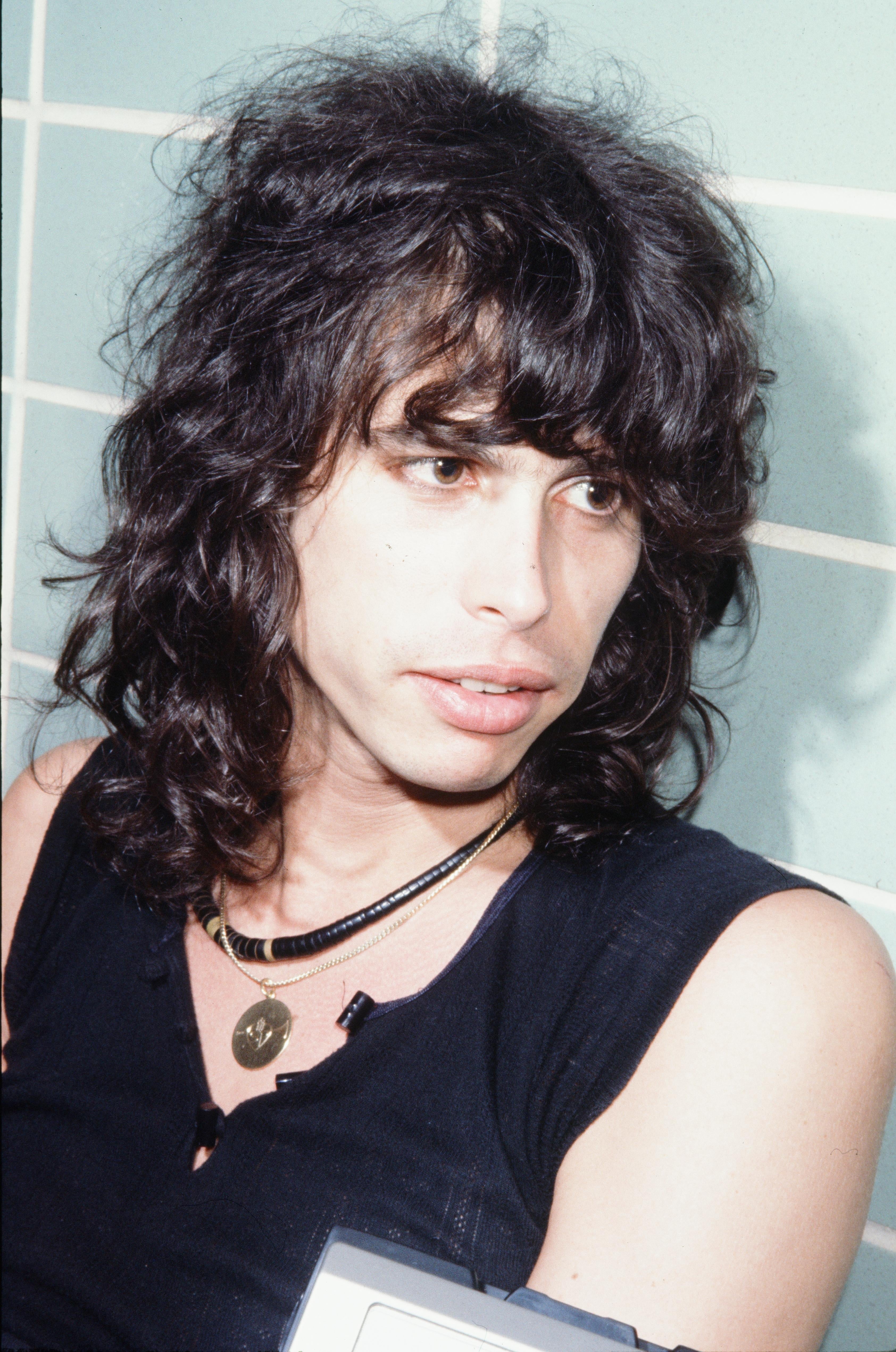 Person with shaggy, shoulder-length hair and layered necklaces, wearing a sleeveless top, gazes to the side against a tiled background