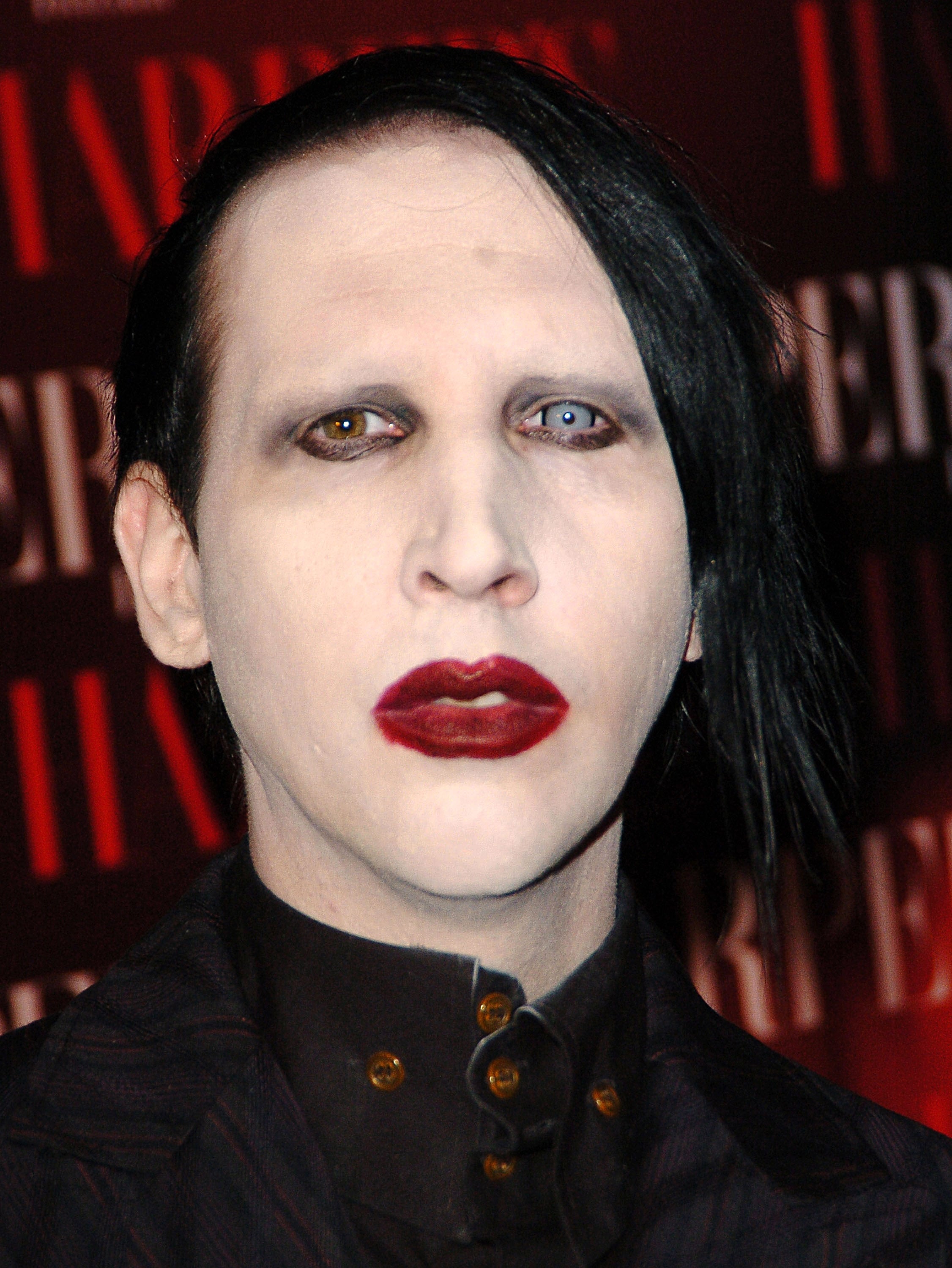 A person on the red carpet wearing a dark, buttoned-up outfit with distinctive makeup and styled hair against a branded backdrop