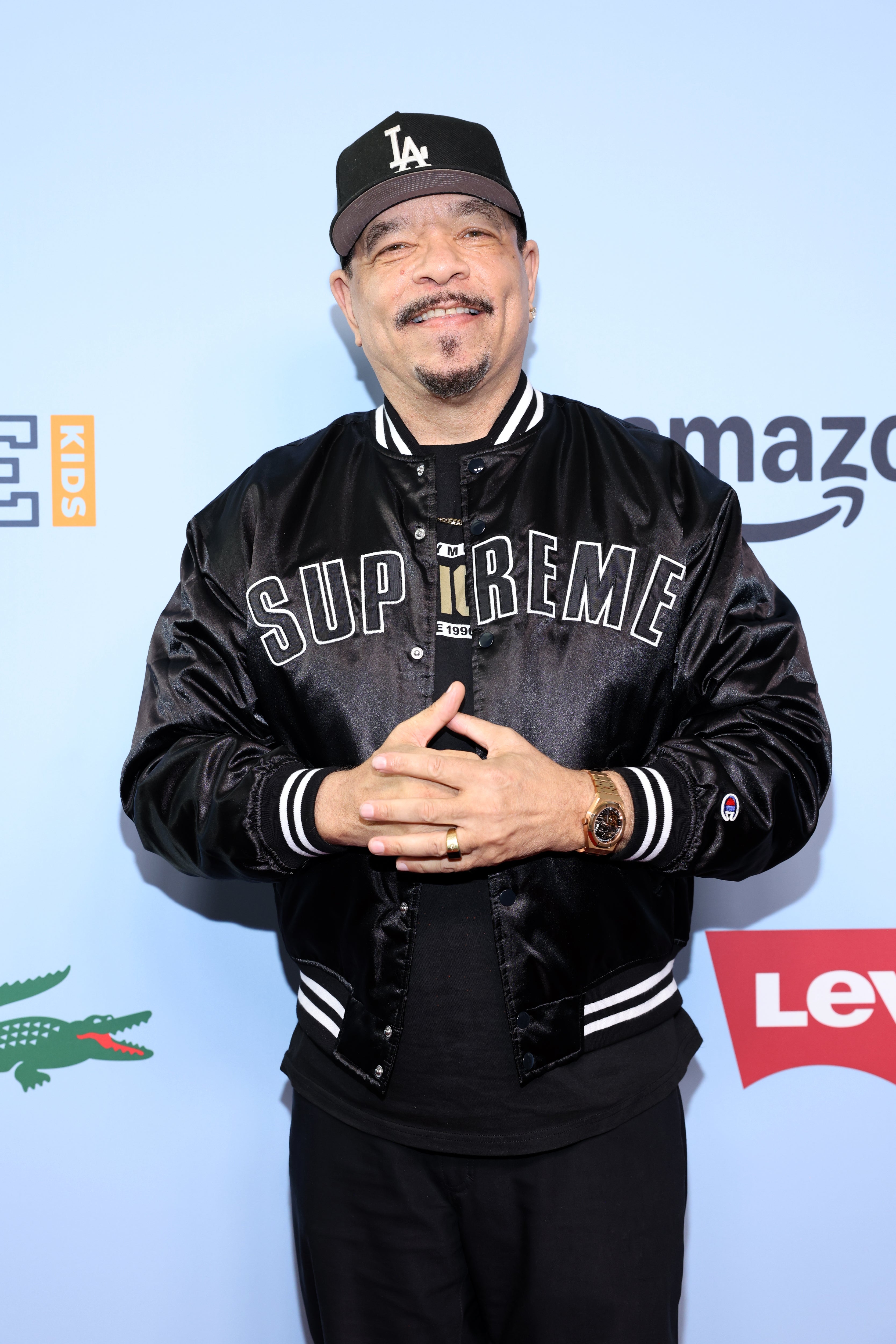 Person in a black baseball cap and a black jacket with bold text on it, smiling casually with hands together, in front of a backdrop with logos