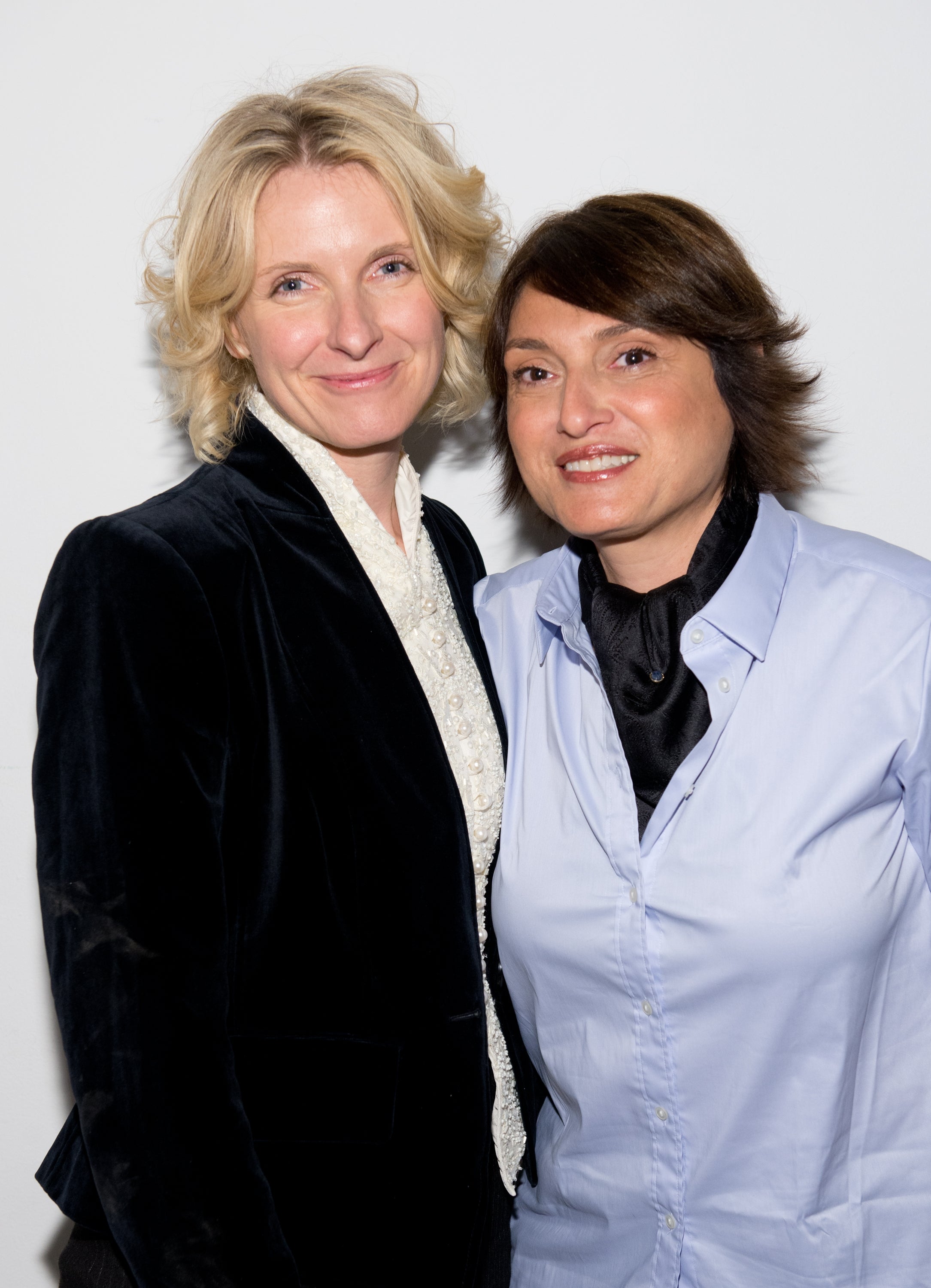 Two people smiling at the camera. One is wearing a velvet jacket and the other in a button-up shirt with a scarf