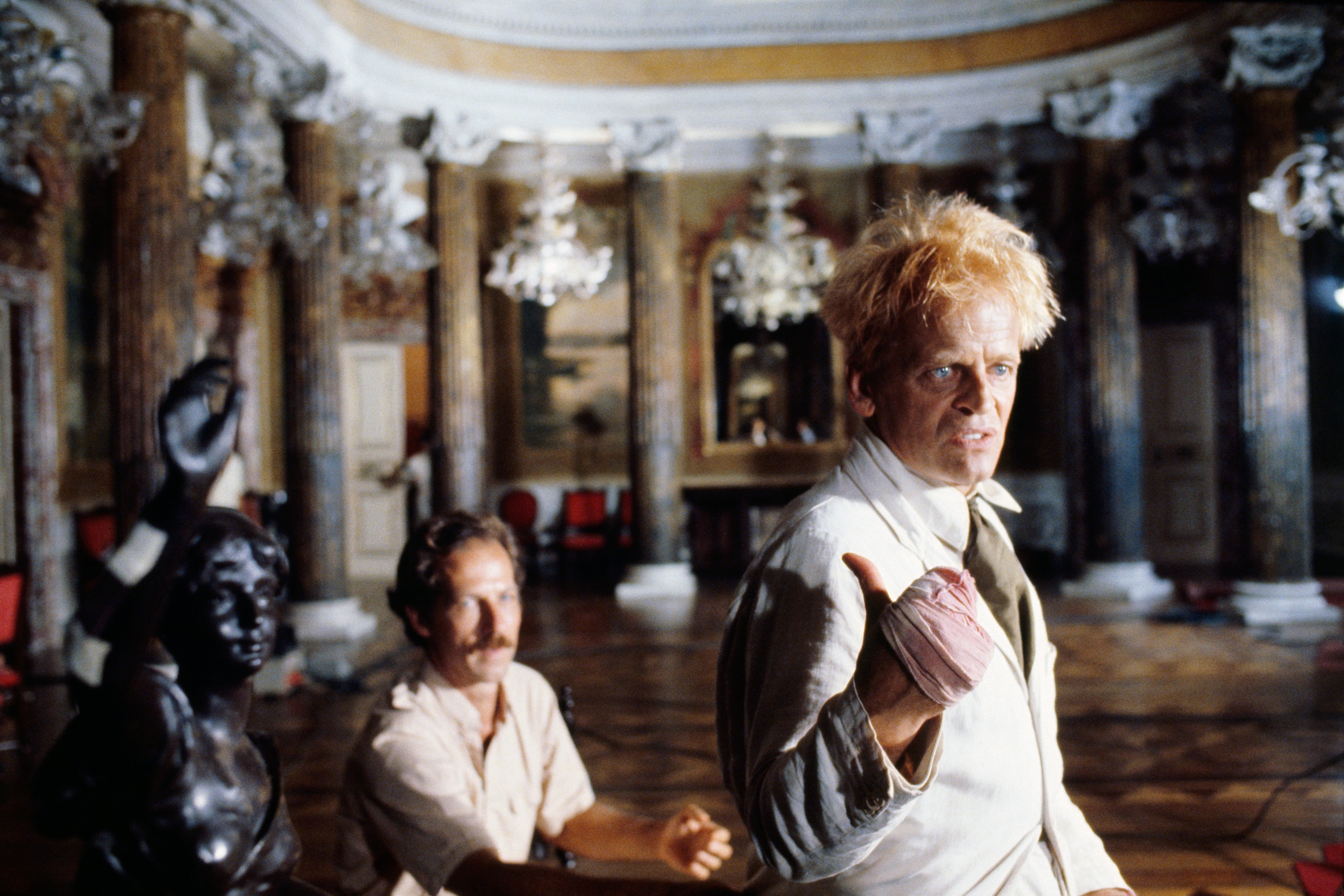 A person with disheveled hair and a bandaged hand poses dramatically in an ornate room with another person in the background
