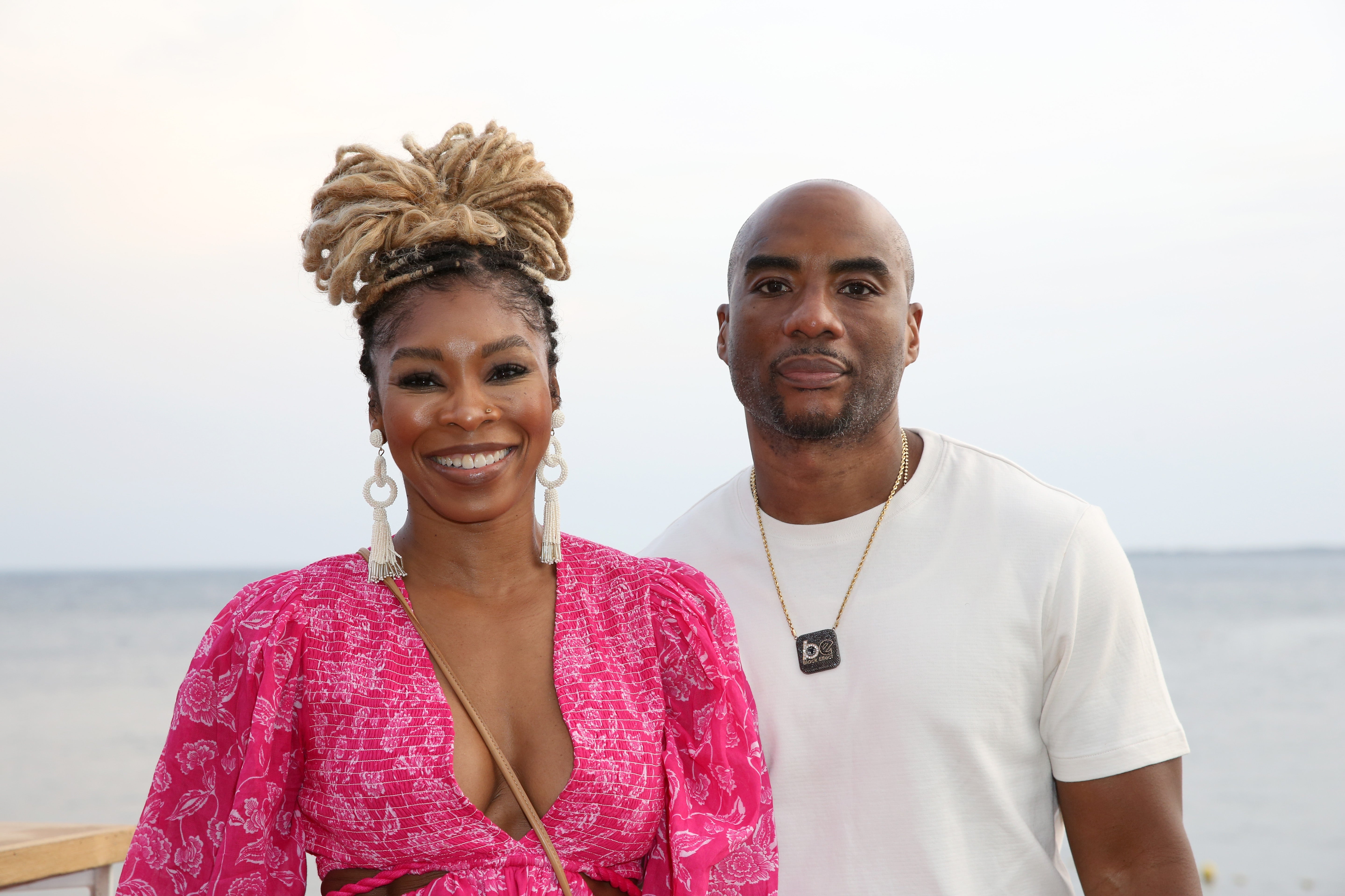 Two individuals smiling at the camera. The woman wears a patterned dress and large earrings, while the man is in a simple t-shirt with a necklace