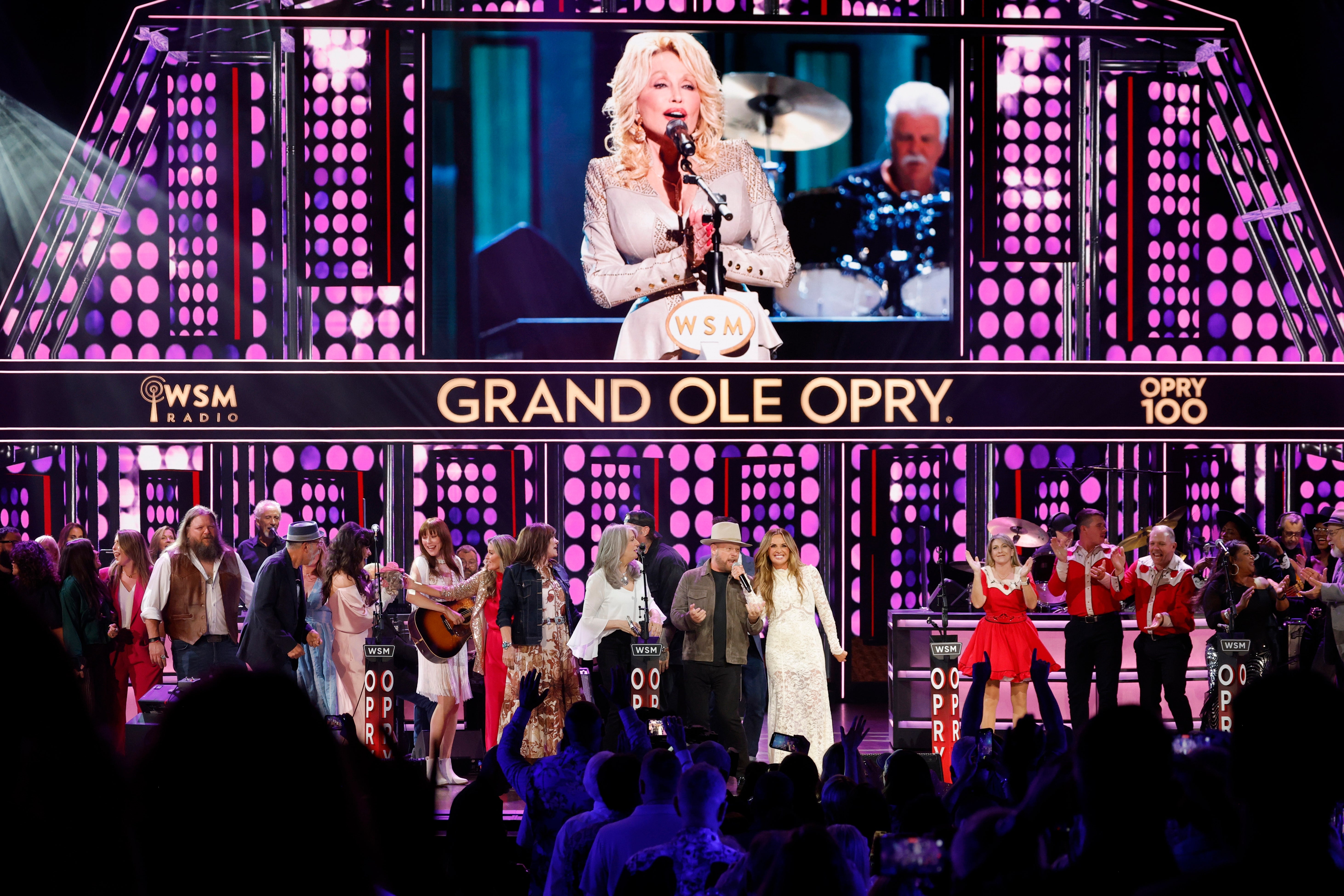 Gruppe med utøvere på scenen på Grand Ole Opry, med en stor skjerm som viser en person som snakker i en mikrofon