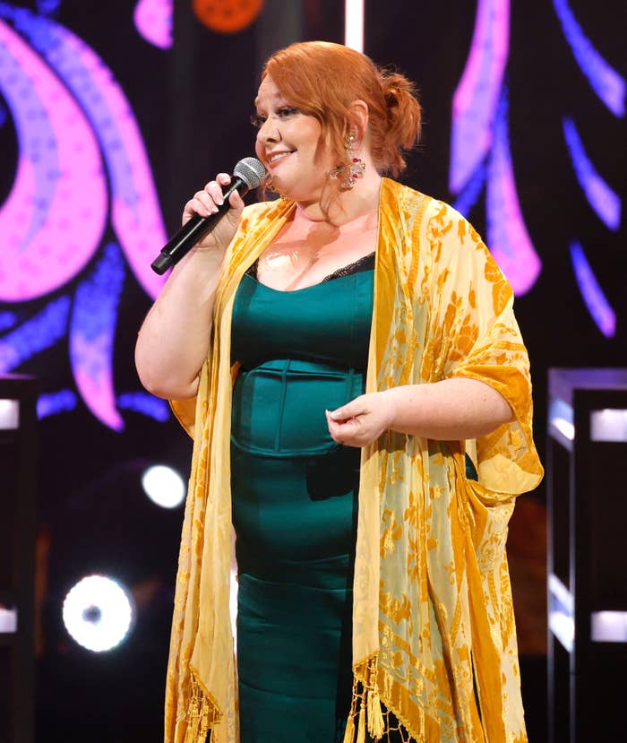 Person on stage holding a microphone, wearing a sleeveless dress and a patterned shawl, performing at a live event