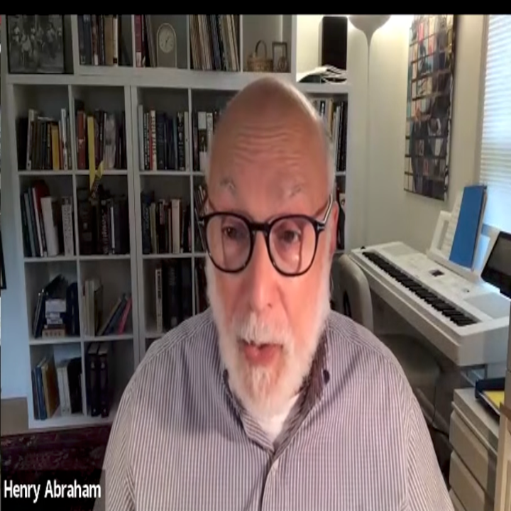 Older adult with glasses and a beard speaks during a video call, set in a home office with bookshelves and a keyboard in the background