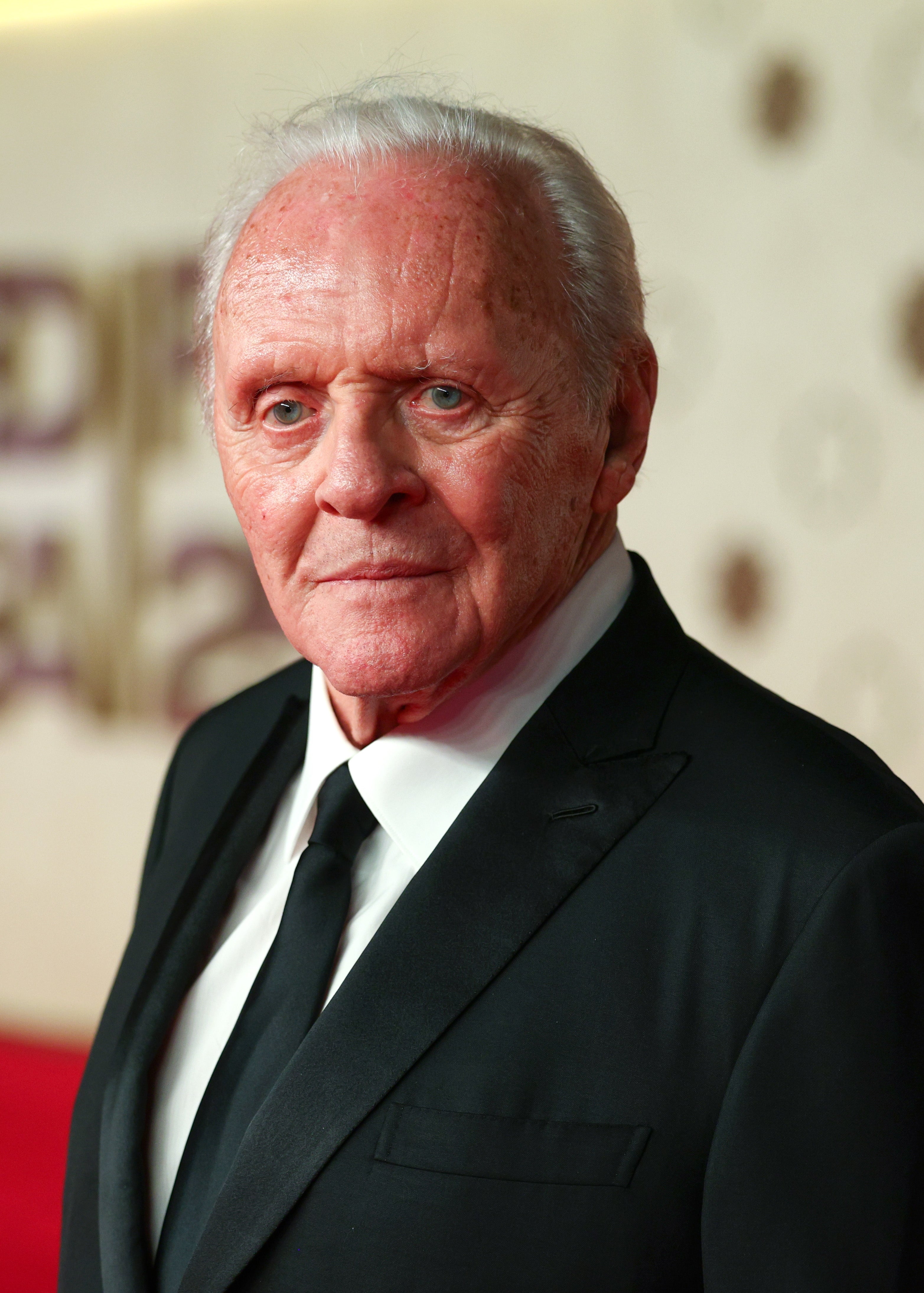 Elderly man in a sharp black suit and tie poses on a red carpet