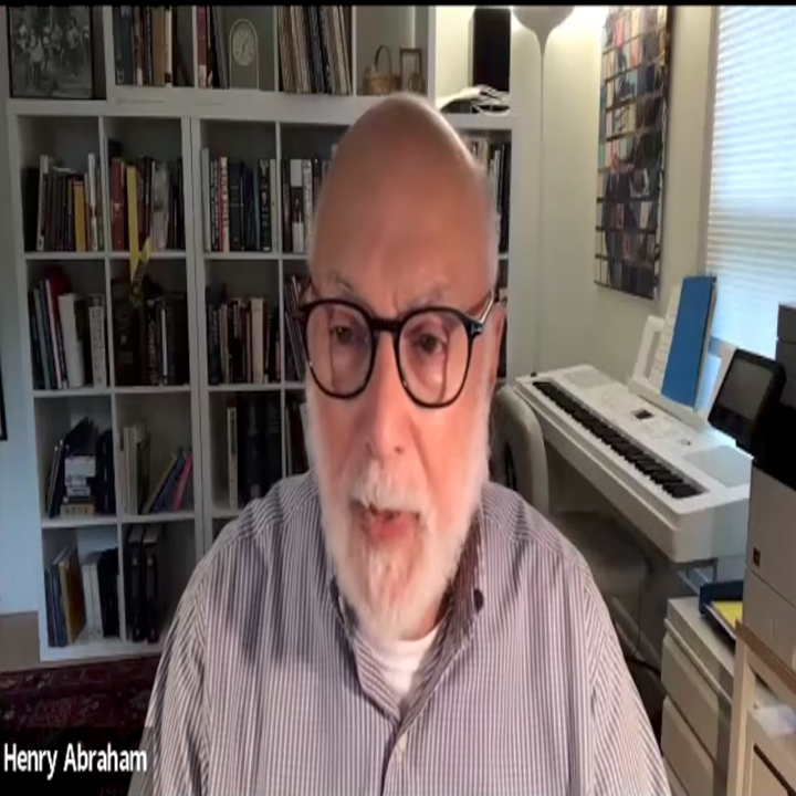 An older man with glasses and a beard is speaking in a room with bookshelves and a keyboard behind him
