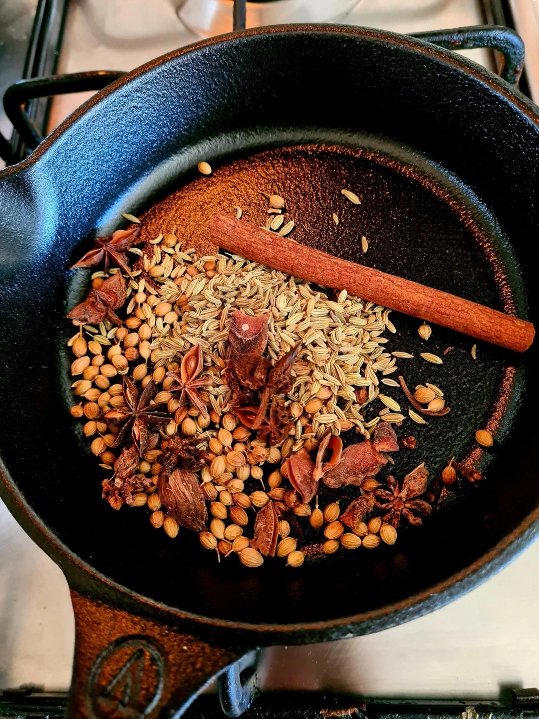 A cast iron pan with various spices including cinnamon, star anise, coriander, and fennel seeds