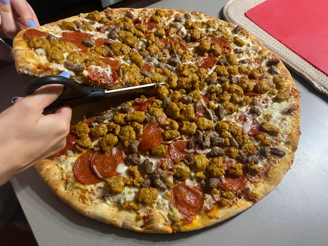 A large pizza topped with pepperoni, sausage, and other toppings is being cut with scissors on a table
