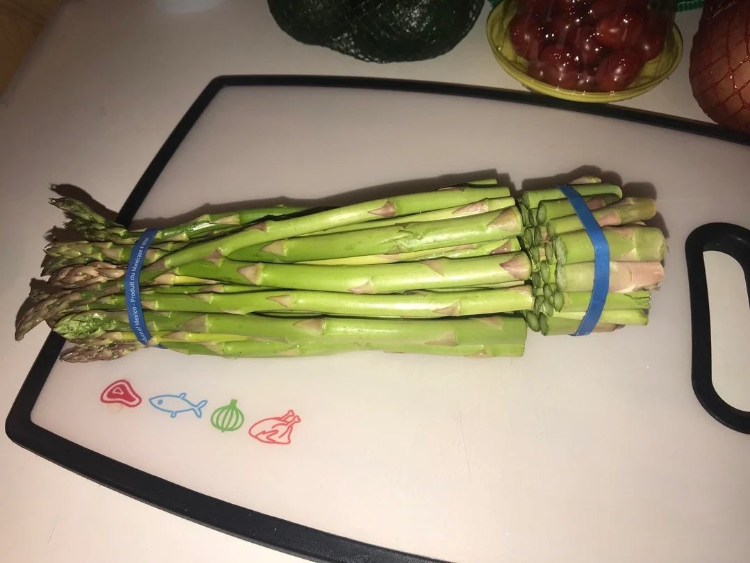 A bundle of fresh asparagus tied with rubber bands on a cutting board, surrounded by avocados, cherry tomatoes, and an onion