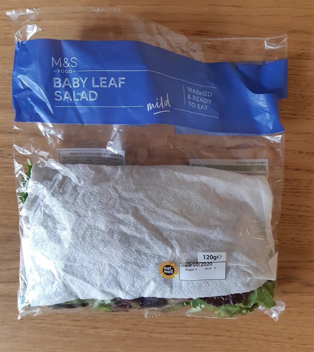 A packaged bag of M&amp;amp;S baby leaf salad, labeled as mild, washed, and ready to eat, placed on a wooden surface