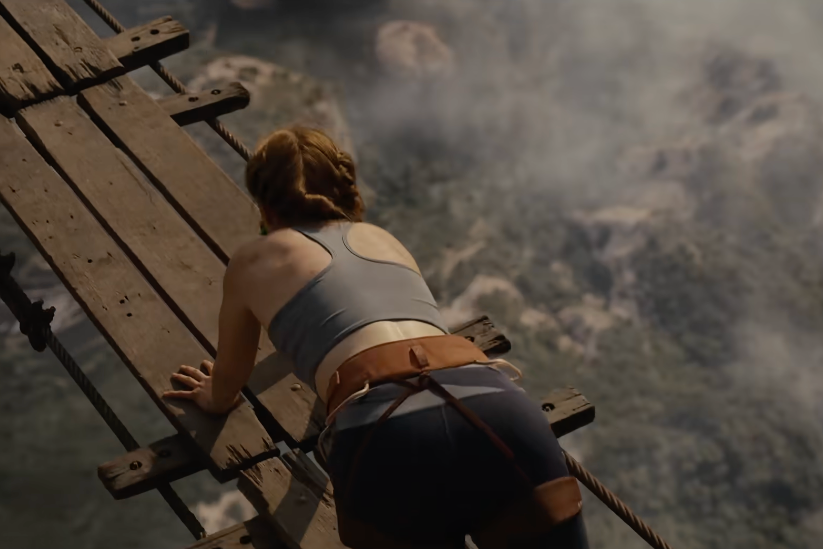 A person in climbing gear is on a wooden bridge over a misty landscape, seemingly performing a daring outdoor adventure
