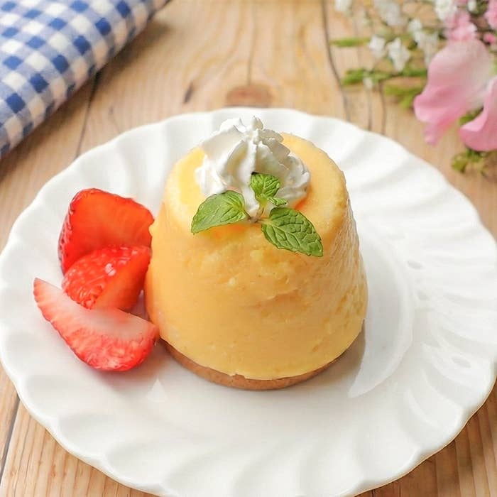 レモンケーキが白い皿にのり、クリームとミント、イチゴがトッピングされています。背景に花とテーブルクロス。