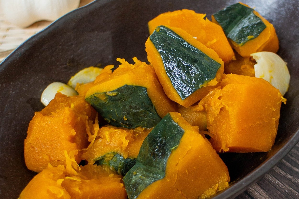 かぼちゃの煮物が黒い皿に盛られた料理。後ろにニンニクが置いてあります。
