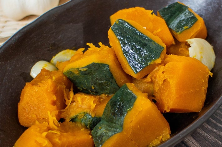 かぼちゃの煮物が黒い皿に盛られた料理。後ろにニンニクが置いてあります。