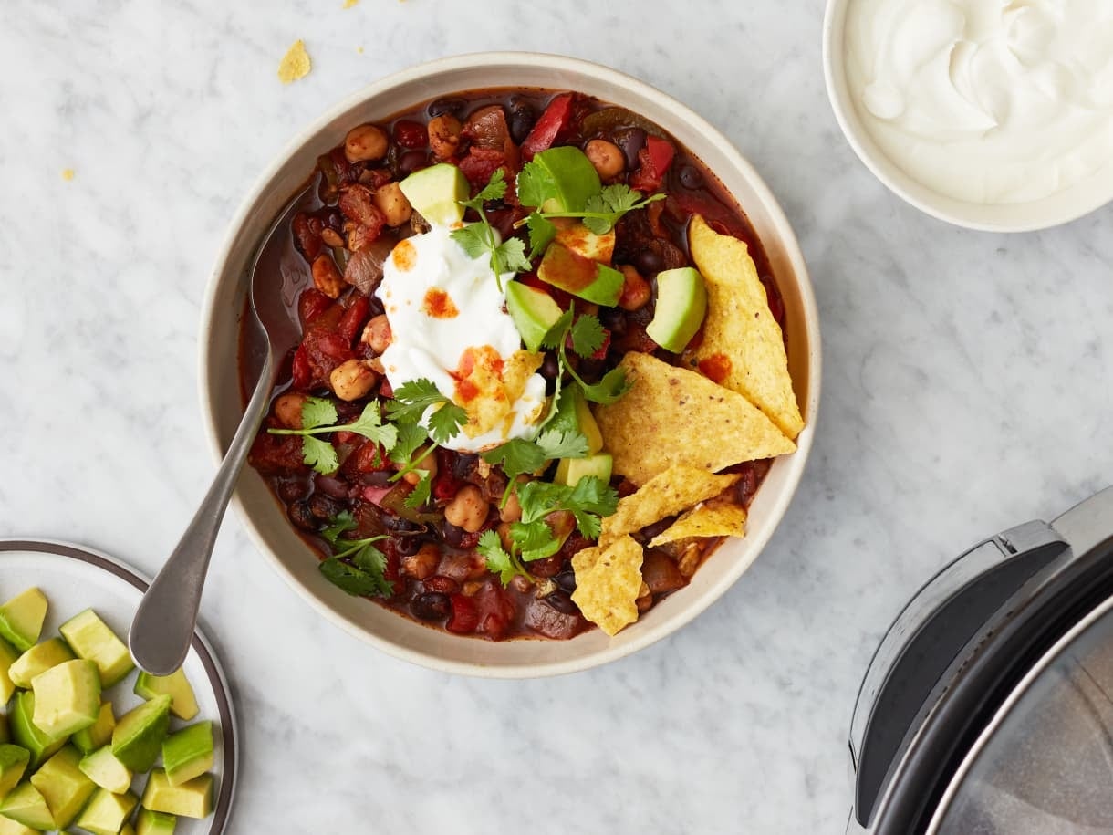 Bowl of chili topped with sour cream, cilantro, and avocado with tortilla chips on the side; avocado chunks and extra sour cream nearby