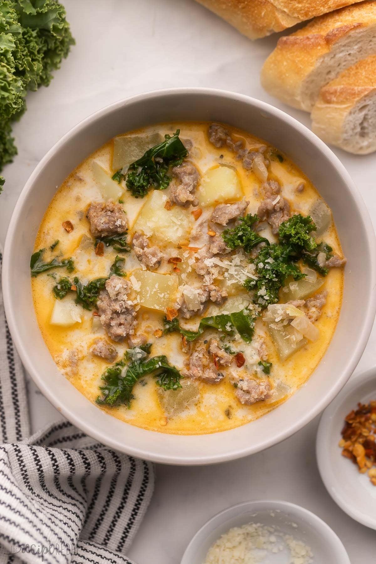 A bowl of creamy soup with sausage, potatoes, kale, and grated cheese, garnished with herbs; bread slices in the background