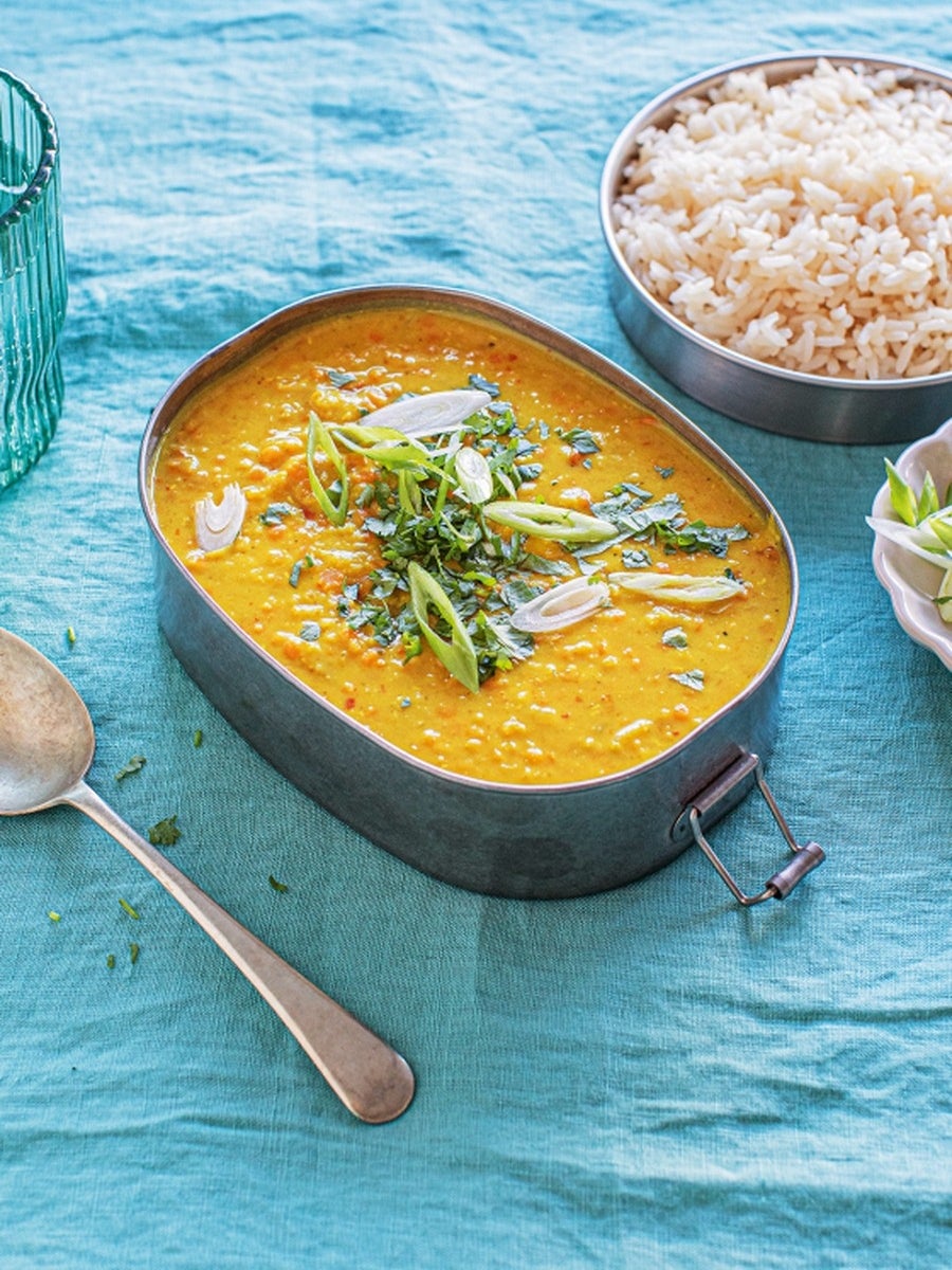 A dish of lentil curry garnished with herbs and green onions, served with a side of rice, on a blue cloth with a glass and spoon nearby
