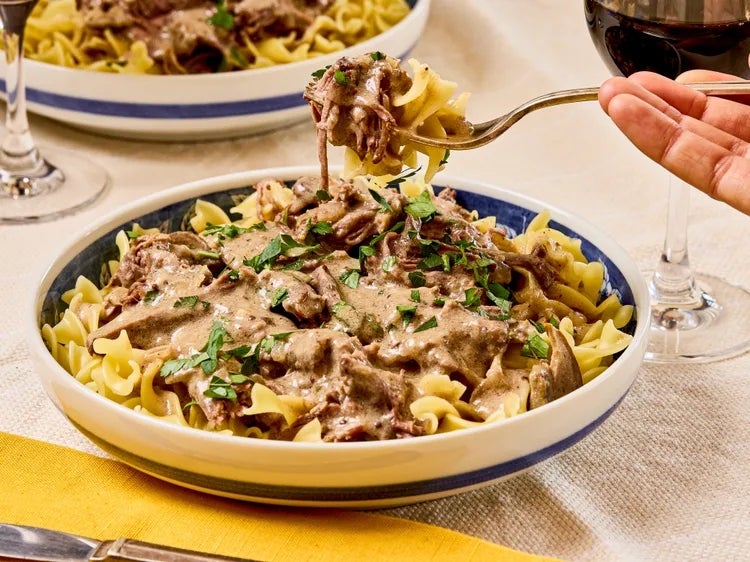 A fork holds a bite of beef stroganoff over a plate of pasta garnished with herbs, next to a glass of red wine