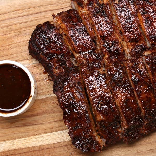 Sliced barbecue ribs on a wooden board with a small bowl of sauce beside them