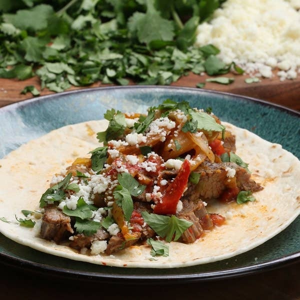 A taco on a plate filled with beef, grilled vegetables, fresh cilantro, and crumbled cheese, with garnishes in the background