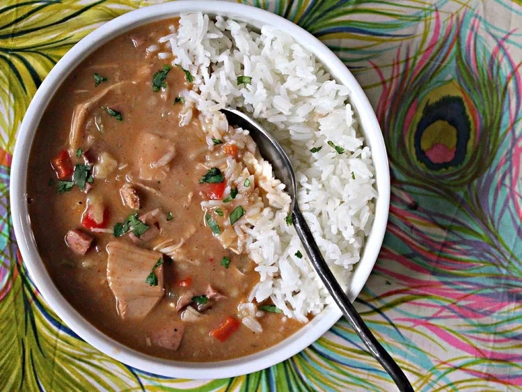 A bowl of rice and creamy stew with vegetables and herbs, placed on a peacock feather-patterned tablecloth. A spoon is in the bowl