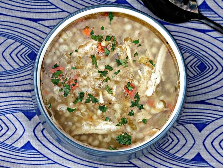 A bowl of soup with visible chicken pieces, small pasta, herbs, and vegetables on a patterned tablecloth