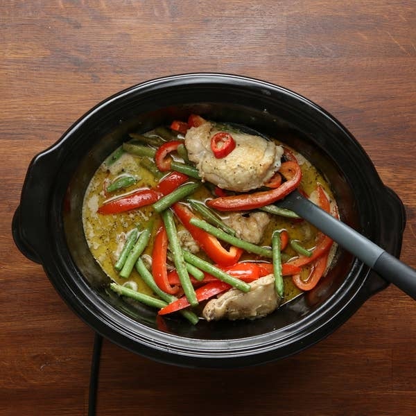 A crockpot filled with a meal of cooked chicken, sliced bell peppers, green beans, and a sauce, with a ladle on the side