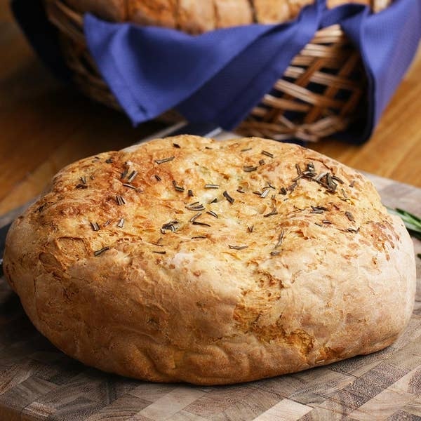 Round loaf of bread with rosemary on a wooden board, partially wrapped basket in the background