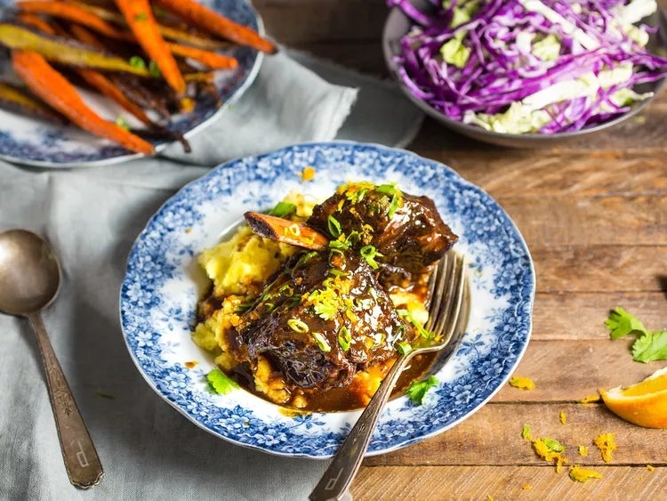 Braised short ribs on mashed potatoes with garnish, served on a floral plate, accompanied by roasted carrots and a slaw with purple cabbage