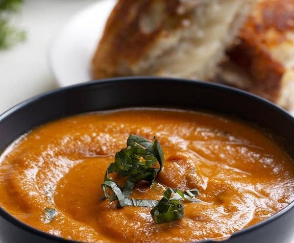 Bowl of creamy tomato soup garnished with basil, served with a piece of bread in the background