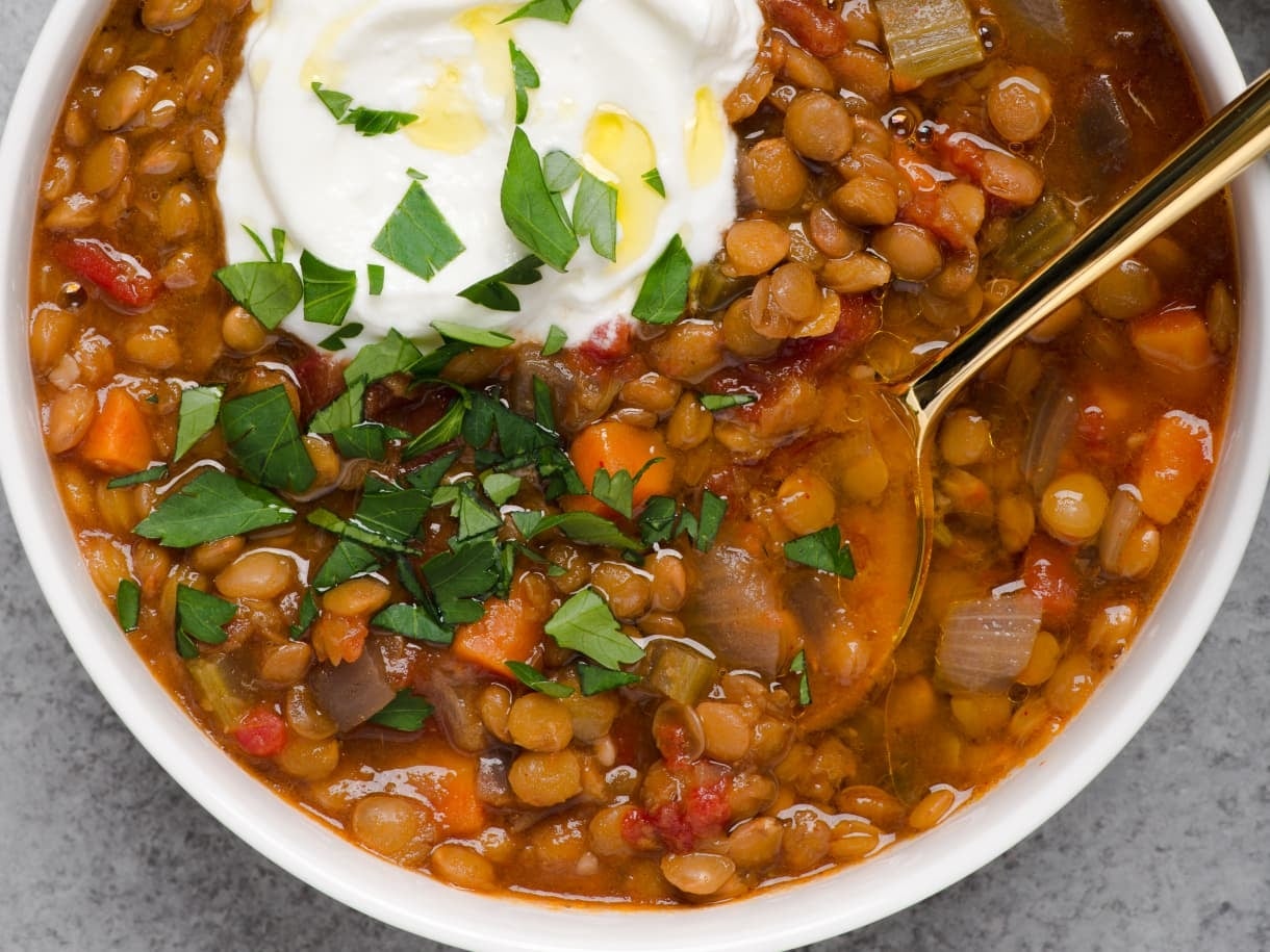 A bowl of lentil soup with chopped vegetables, garnished with a dollop of cream and fresh herbs, with a golden spoon