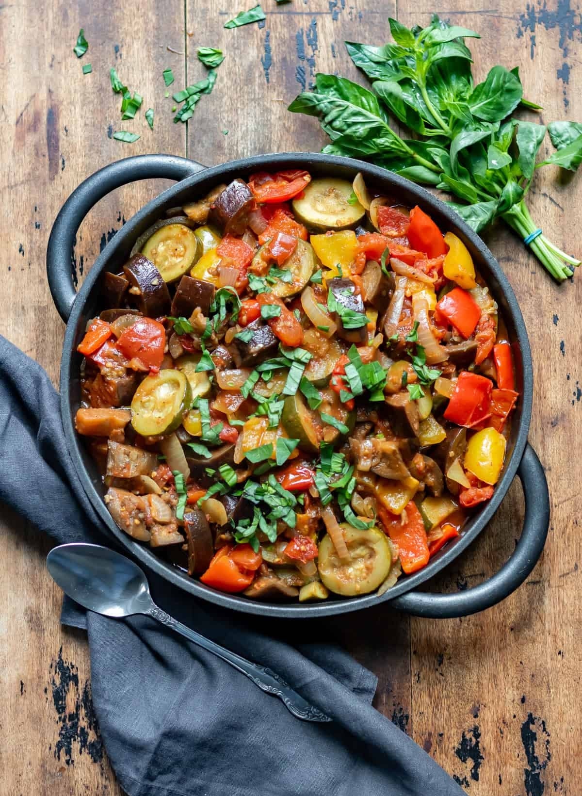 A rustic skillet of ratatouille with sliced zucchini, eggplant, bell peppers, and tomatoes, garnished with fresh basil beside a wooden table