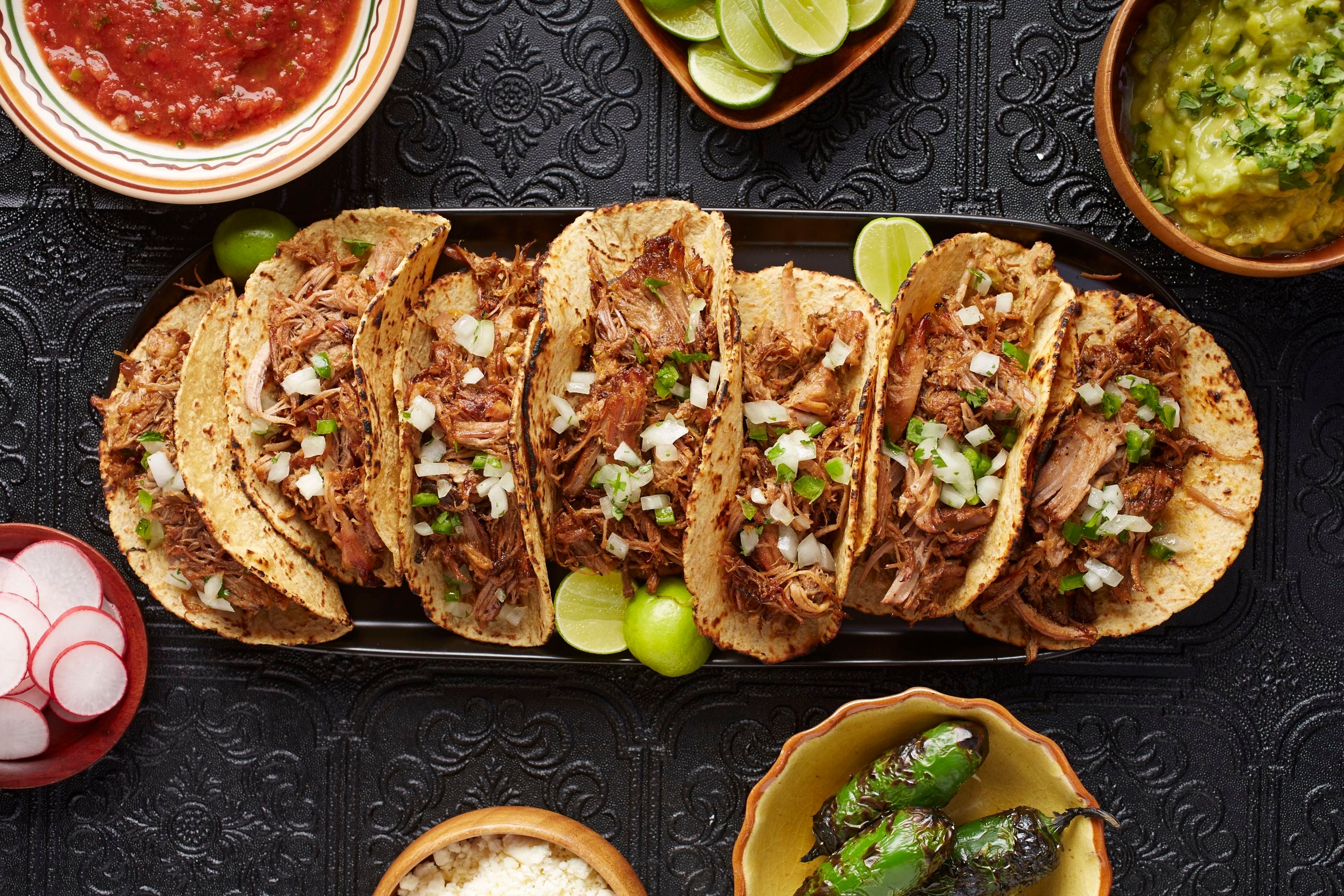 Plate of tacos filled with shredded meat and onions, surrounded by bowls of salsa, lime, guacamole, radishes, cheese, and grilled jalapeños