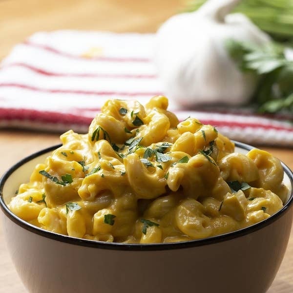 A bowl of creamy macaroni and cheese garnished with chopped herbs, placed on a wooden table with a garlic bulb and towel in the background